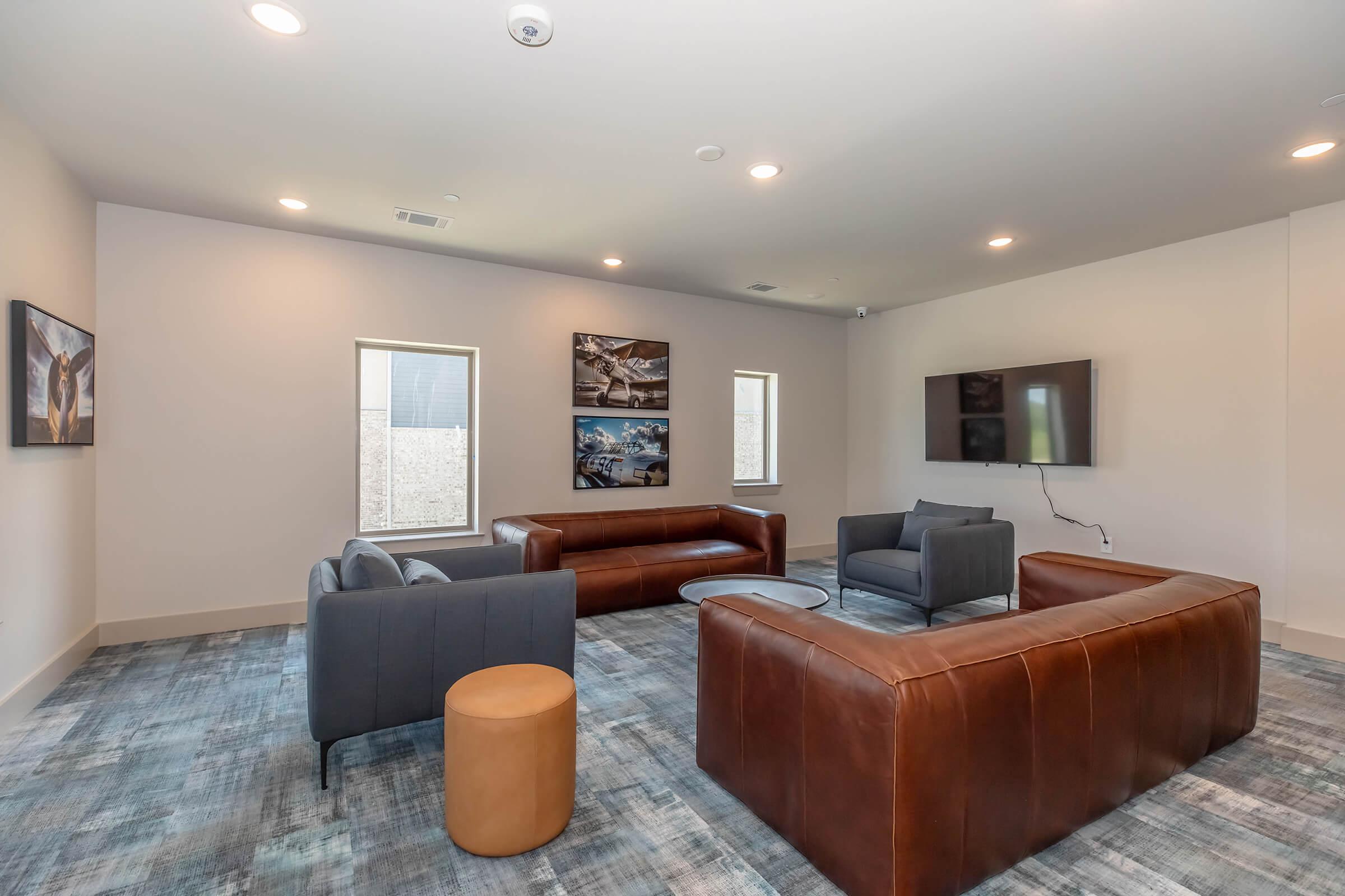 A modern lounge area featuring two brown leather sofas, a gray armchair, and a round coffee table. The walls are adorned with framed photos, and there’s a flat-screen TV mounted on the wall. Natural light pours in through two windows, and the floor is covered with a textured carpet.