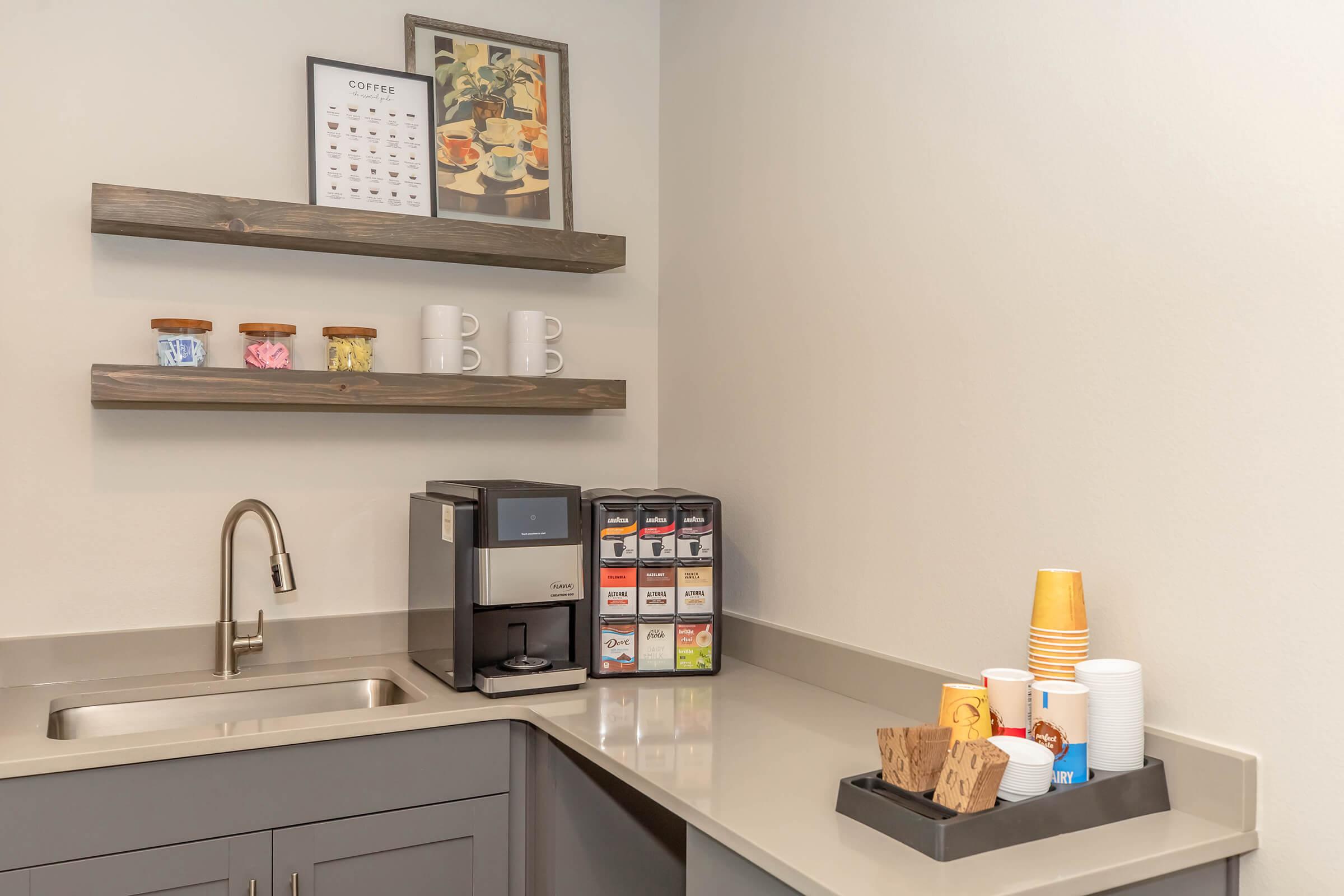 A modern coffee station featuring a coffee machine, a variety of tea and coffee packets, colorful mugs, and jars with snacks or condiments. The setup includes a sink and minimalist shelving with a coffee-related poster, all arranged in a clean, contemporary kitchen space.