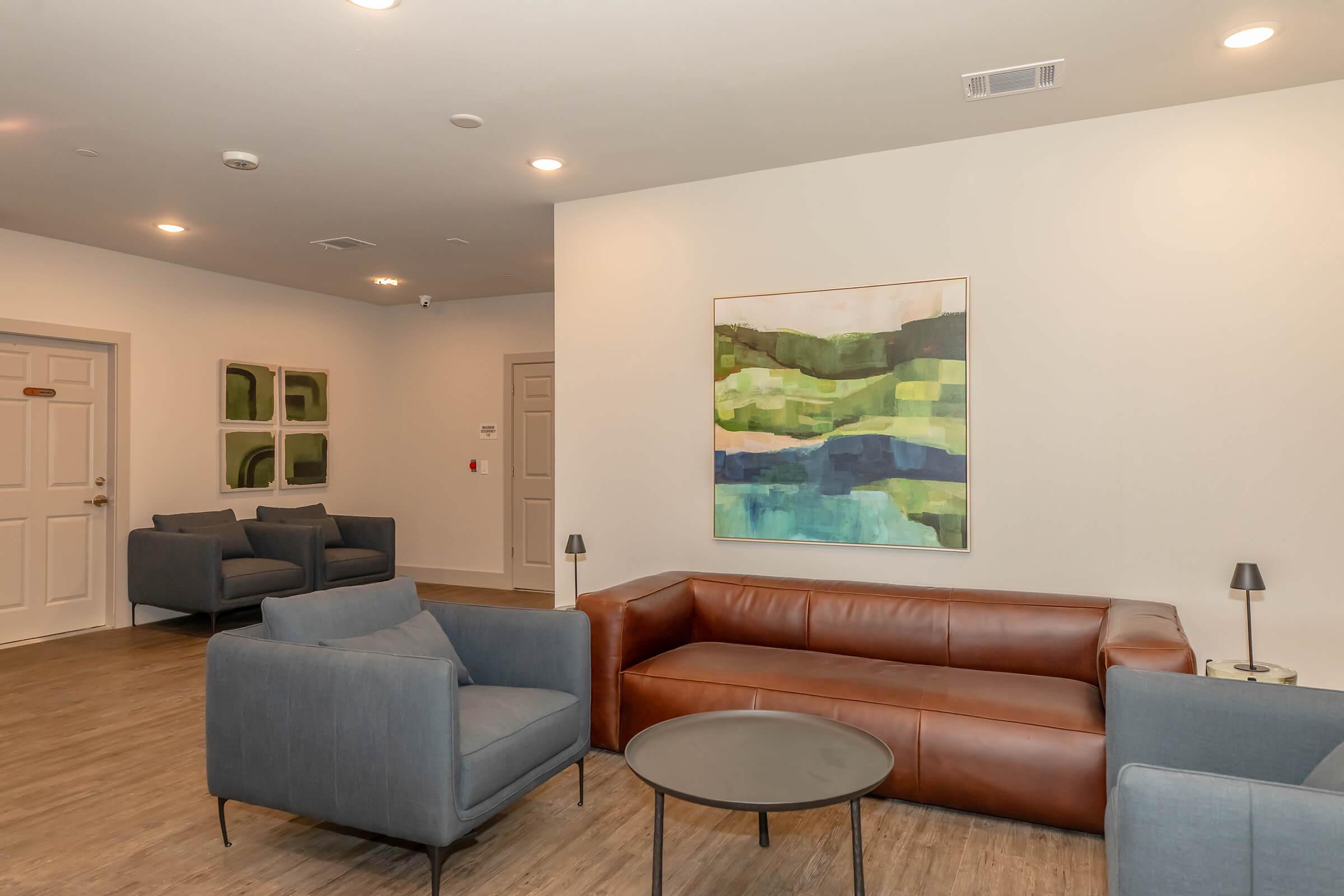 A modern lounge area featuring a brown leather sofa, two gray armchairs, and a round black table. The walls are decorated with abstract art in green and blue tones, and there are two additional green artwork pieces on the wall. The flooring is wooden, creating a contemporary and inviting atmosphere.