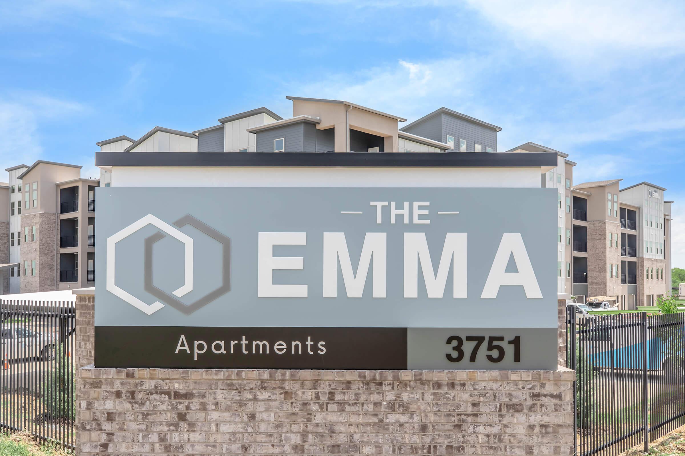Signage for "The Emma" Apartments, prominently displayed in a modern style. The sign features the name "THE EMMA" in large letters, along with the word "Apartments" below. The building in the background has multiple stories, showcasing a contemporary architectural design, with a clear blue sky above.