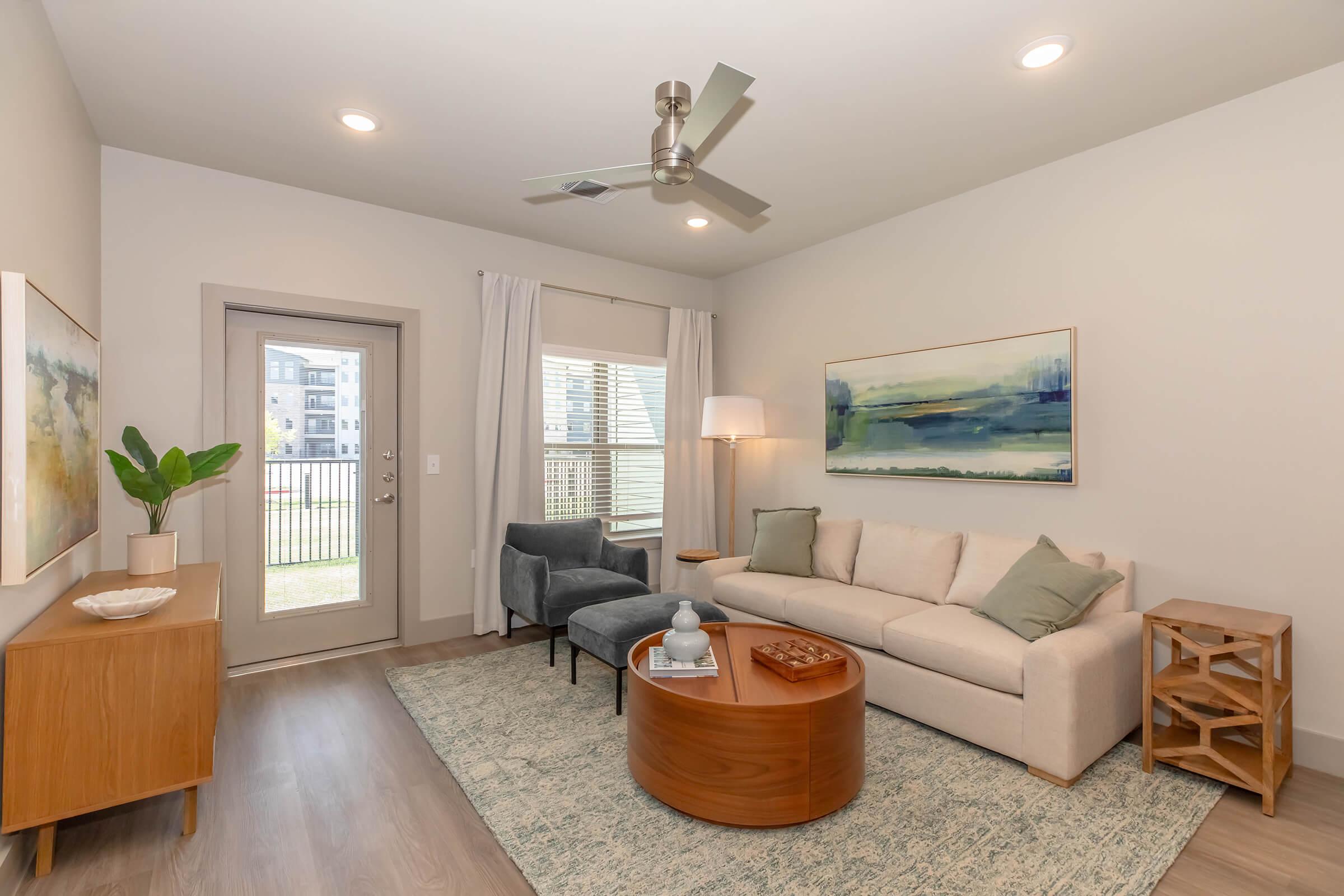 A modern living room featuring a light-colored sofa, a gray accent chair, and a wooden coffee table. There is a decorative plant and abstract wall art. The room is bright with large windows, and a ceiling fan is installed. Natural light streams in, creating a welcoming atmosphere.