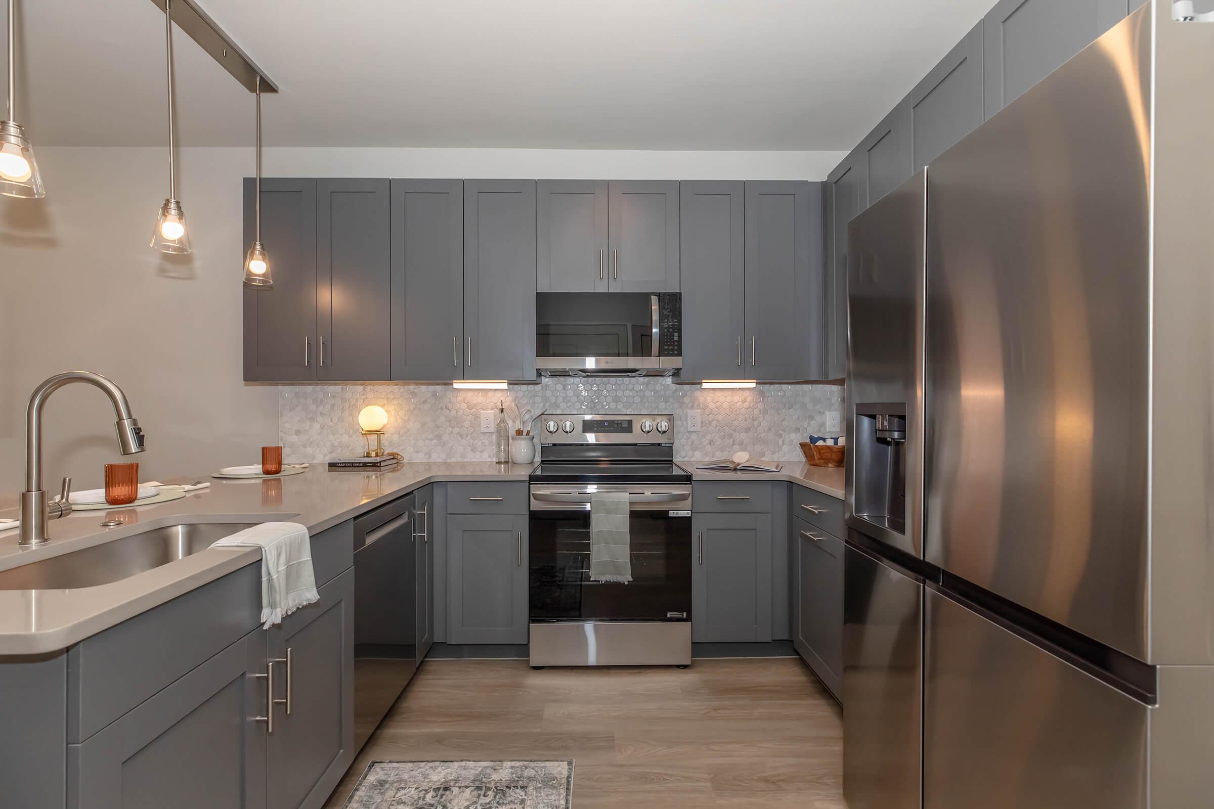 Modern kitchen featuring gray cabinetry, stainless steel appliances, and a marble backsplash. The space includes a large refrigerator, oven, and a sleek countertop with a sink. Pendant lights hang above, creating a warm ambiance. A small decorative item is on the counter, enhancing the contemporary style.