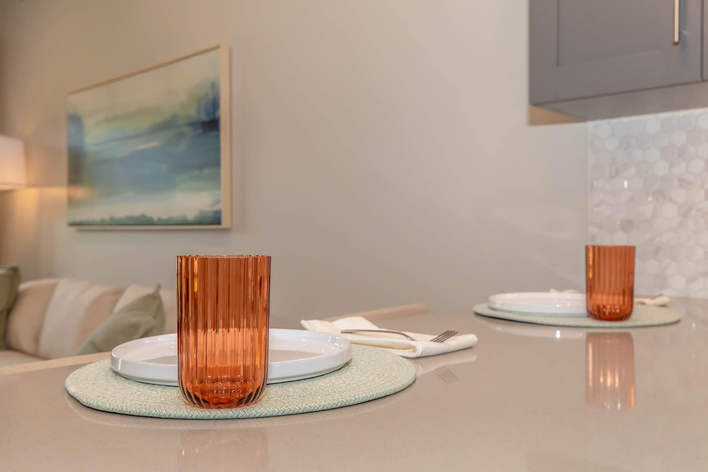 A modern dining setup featuring two white plates on light-colored placemats, complemented by an orange glass on the counter. Soft lighting and an abstract painting in the background add a cozy ambiance to the space.