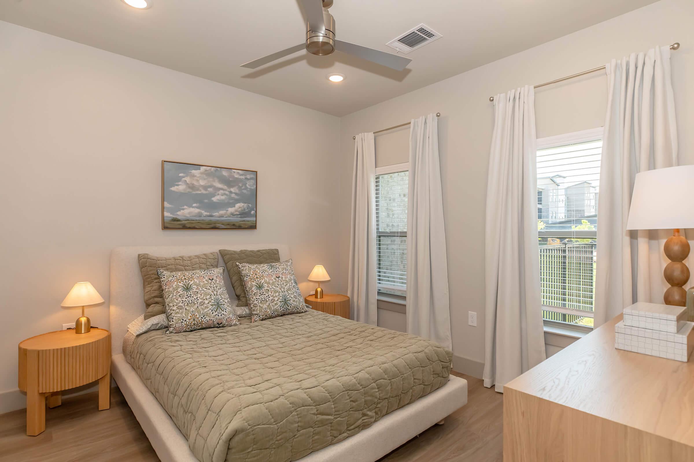 A cozy bedroom featuring a queen-sized bed with a light gray upholstered headboard and decorative pillows. There are two wooden nightstands with lamps on either side of the bed. A large window with sheer curtains lets in natural light, and a painting of clouds hangs on the wall. A wooden dresser is also present.