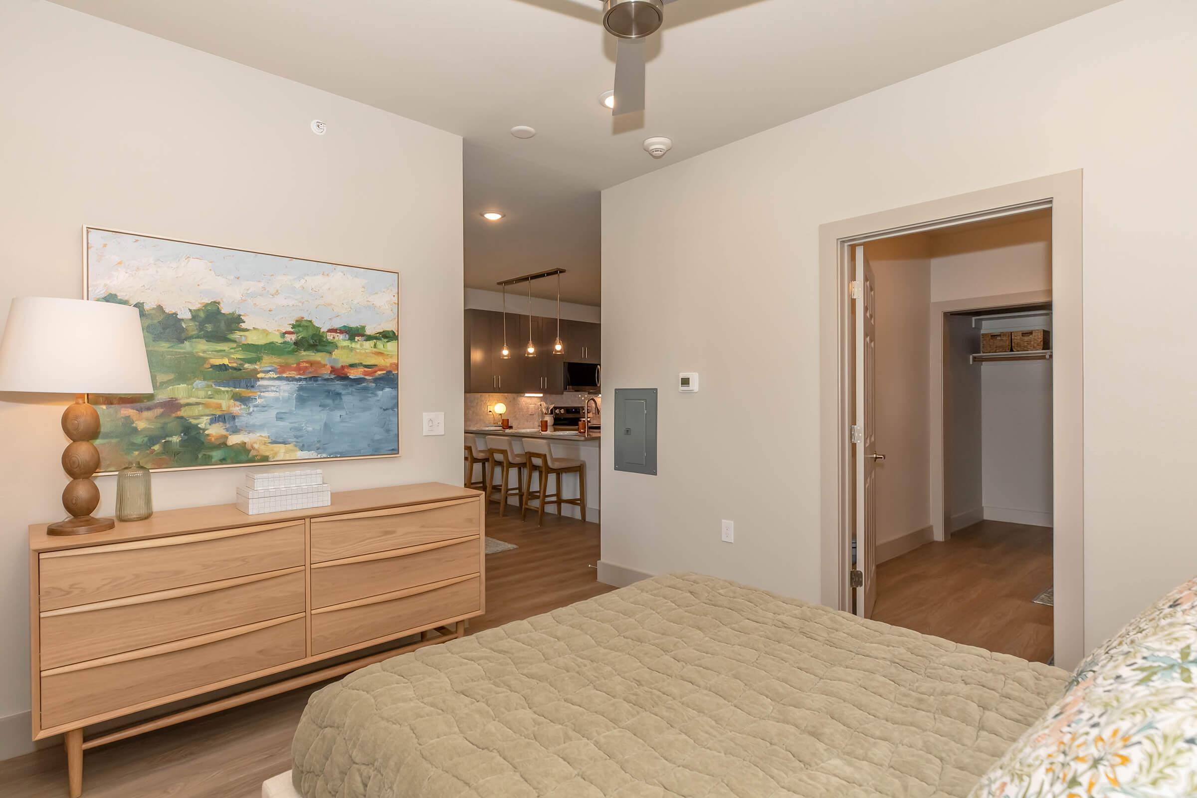 A modern bedroom featuring a bed with a green comforter, a wooden dresser with a lamp, and a colorful landscape painting on the wall. A doorway leads to a closet, while the background reveals an open kitchen area with bar seating and contemporary lighting. The decor is light and inviting.
