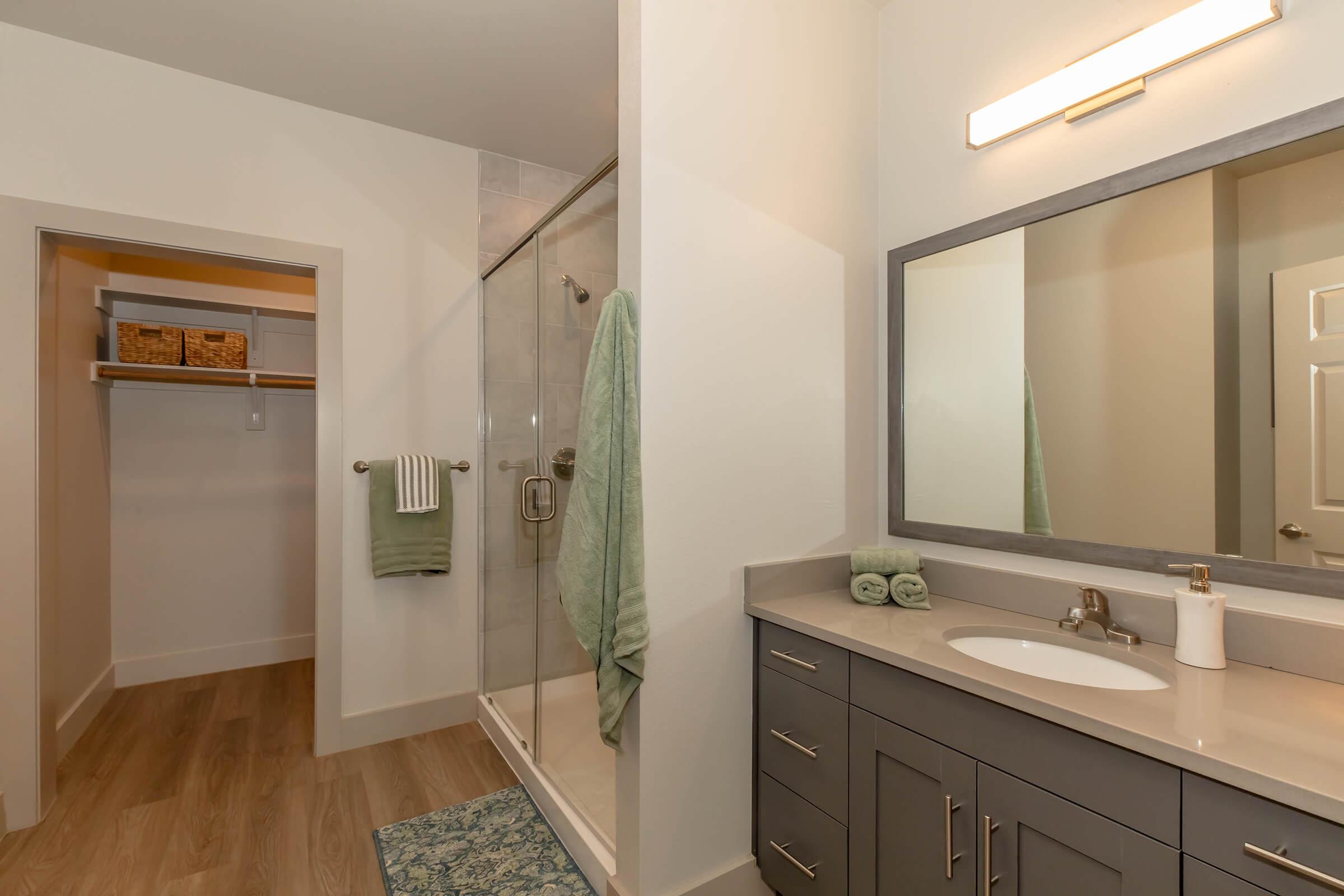A modern bathroom featuring a glass shower, light gray cabinetry with a sink, a large mirror, and a towel rack. There are green towels displayed, and a neatly organized closet space is visible in the background. The flooring is wooden, adding a warm touch to the contemporary design.