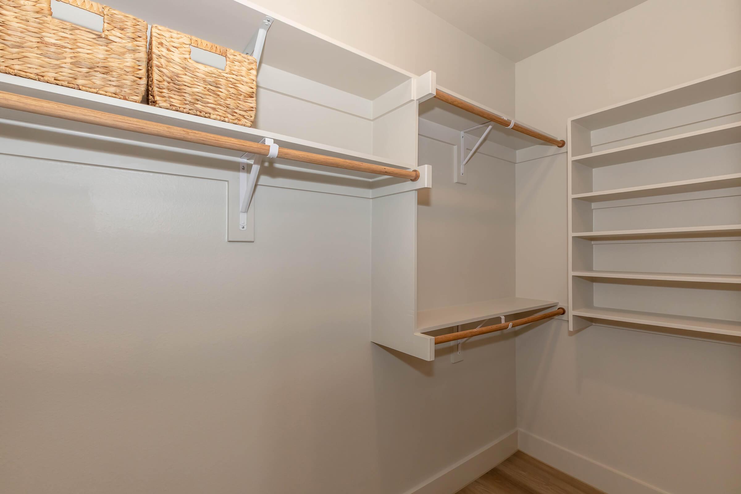 A tidy walk-in closet featuring white shelves, wooden hanging rods, and two woven baskets on top. The layout includes open space for storing clothes and accessories, with organized shelving along the walls and a neutral color scheme.
