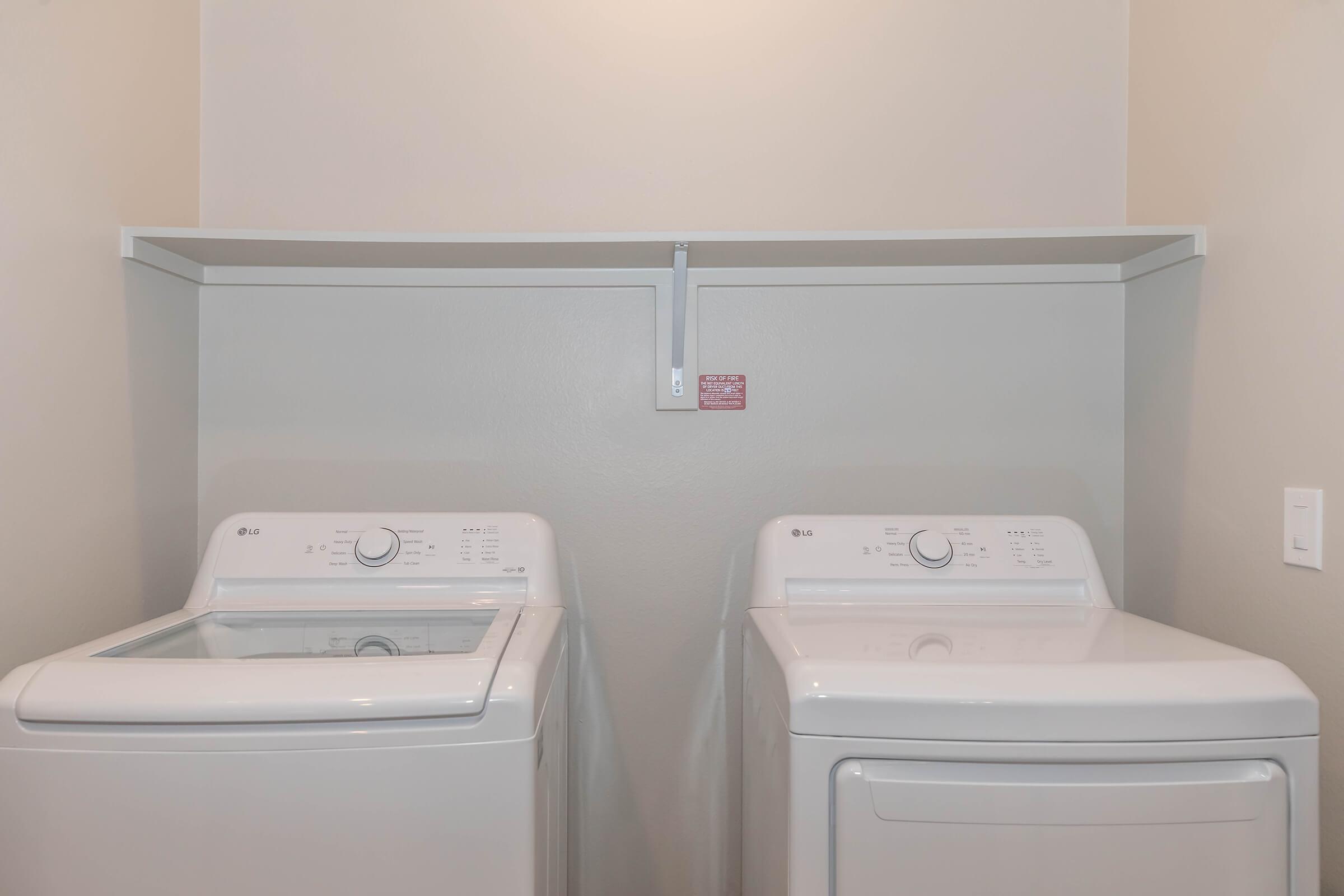 A laundry room featuring a side-by-side washing machine and dryer in white. Above them is a simple shelf mounted on the wall. The walls are painted a neutral color, contributing to a clean, modern look. A notice on the wall is partially visible behind the appliances.