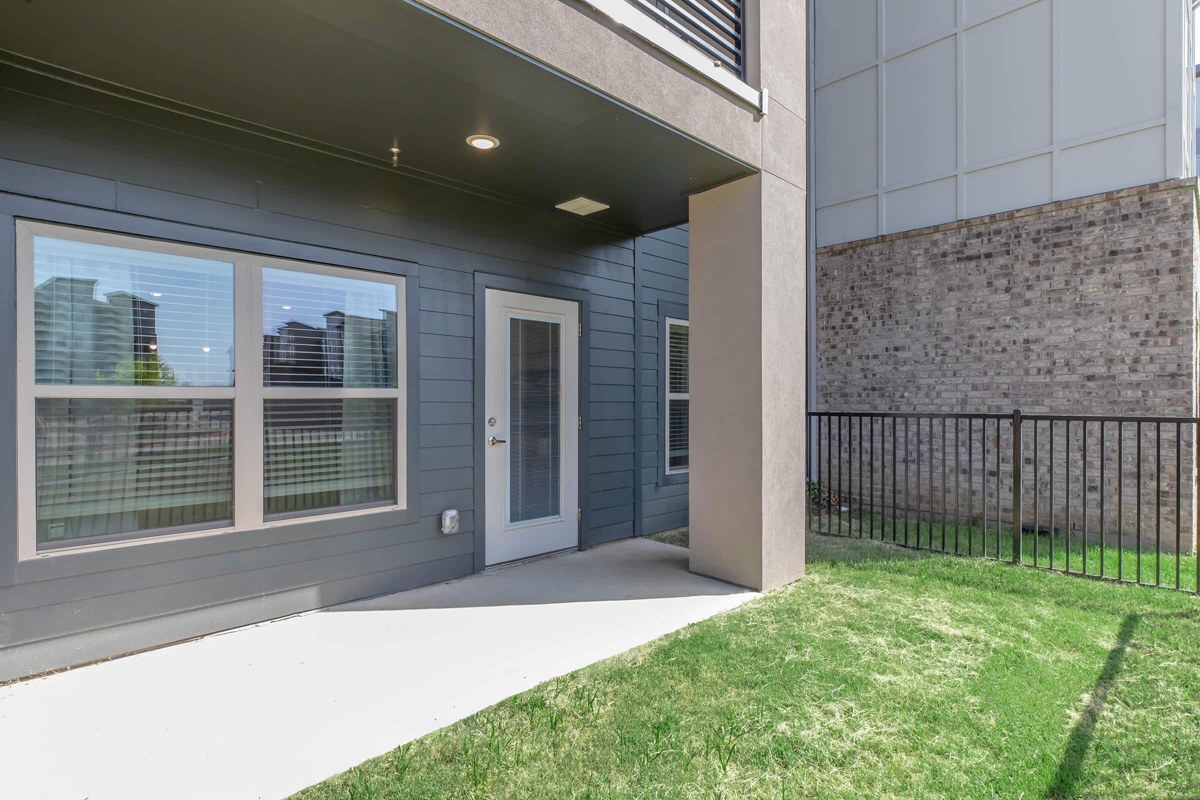 A modern patio area featuring a door leading into a building, flanked by large windows with blinds. The exterior is gray with textured walls, and there's a small, well-maintained lawn space adjacent to a decorative fence. The background shows a more urban environment with nearby buildings.