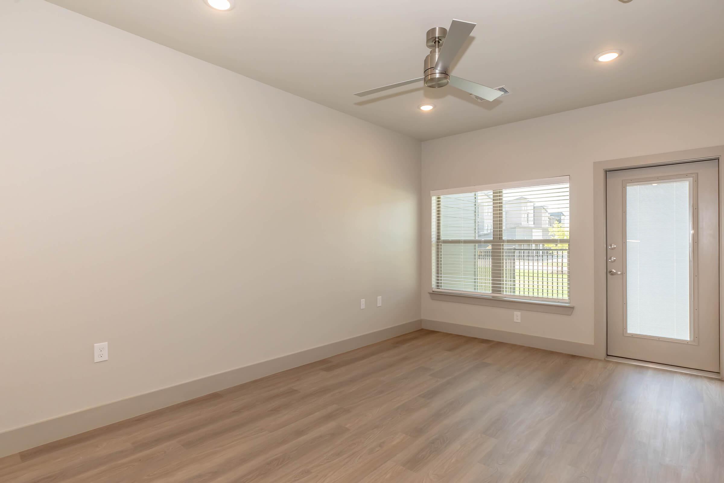 A minimalist room with light gray walls and a ceiling fan. The space features a large window with blinds, allowing natural light to enter. There is a modern glass door on one side, and the floor is made of light-colored wood. The overall atmosphere is bright and open, suitable for various purposes.