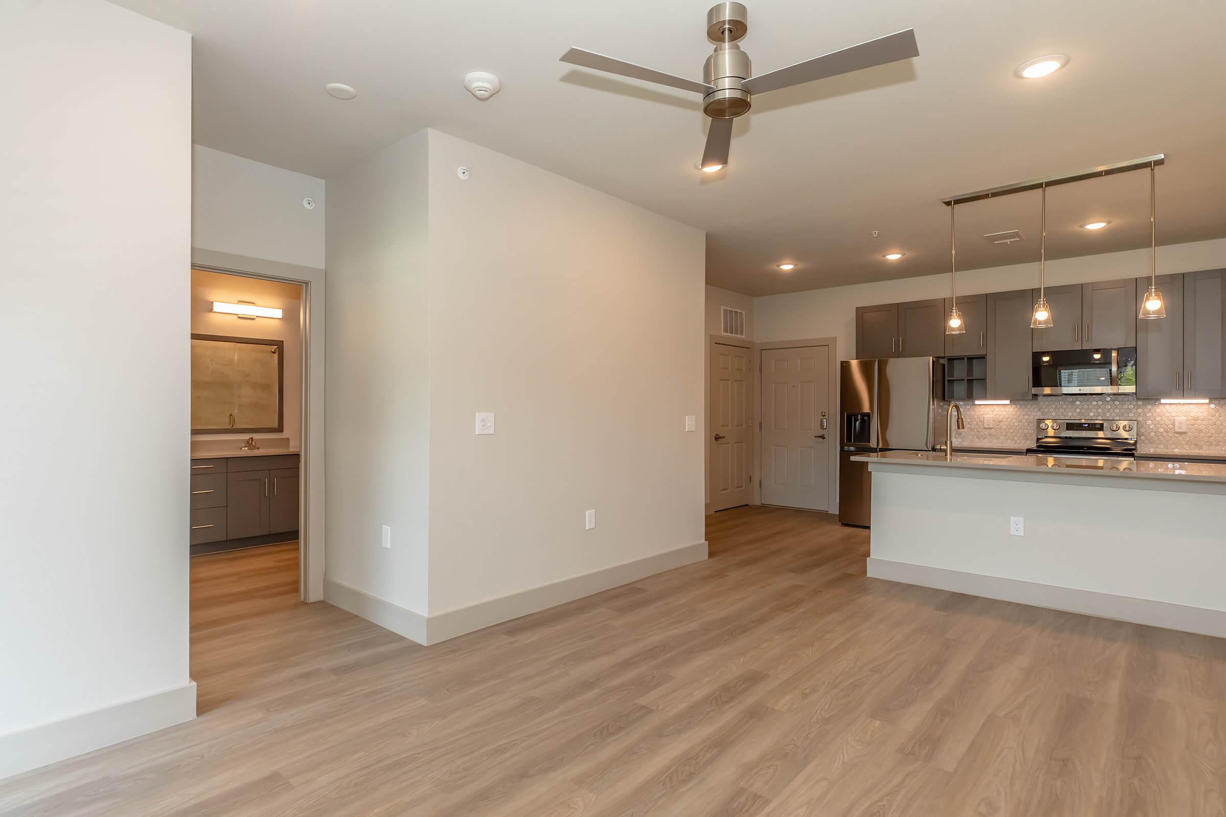 Spacious modern apartment interior featuring an open layout with hardwood-style flooring. The room includes a ceiling fan, contemporary light fixtures, and a view of the kitchen equipped with stainless steel appliances. There is a doorway leading to a bathroom and a neutral color palette throughout.