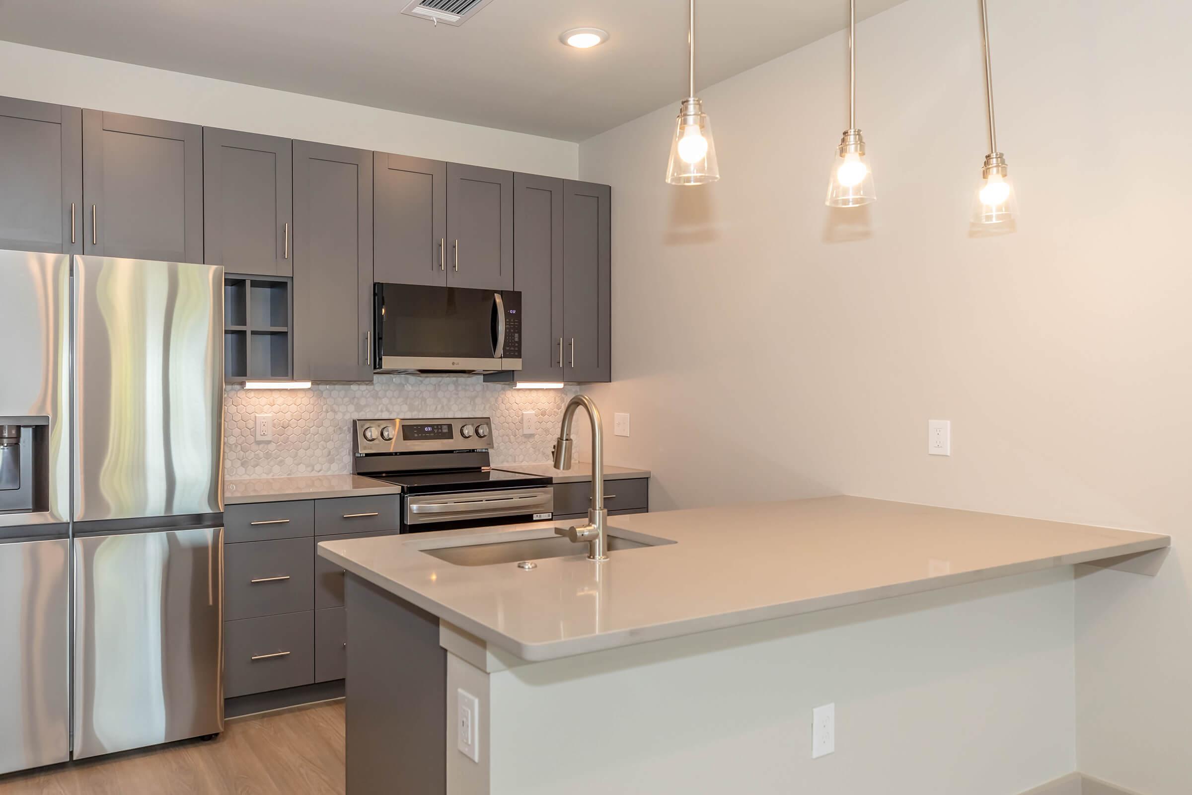 Modern kitchen featuring sleek gray cabinets, stainless steel appliances including a refrigerator and microwave, and a countertop with a built-in sink. Pendant lights hang above the island, creating a bright and inviting atmosphere. The decor is minimalist and contemporary.