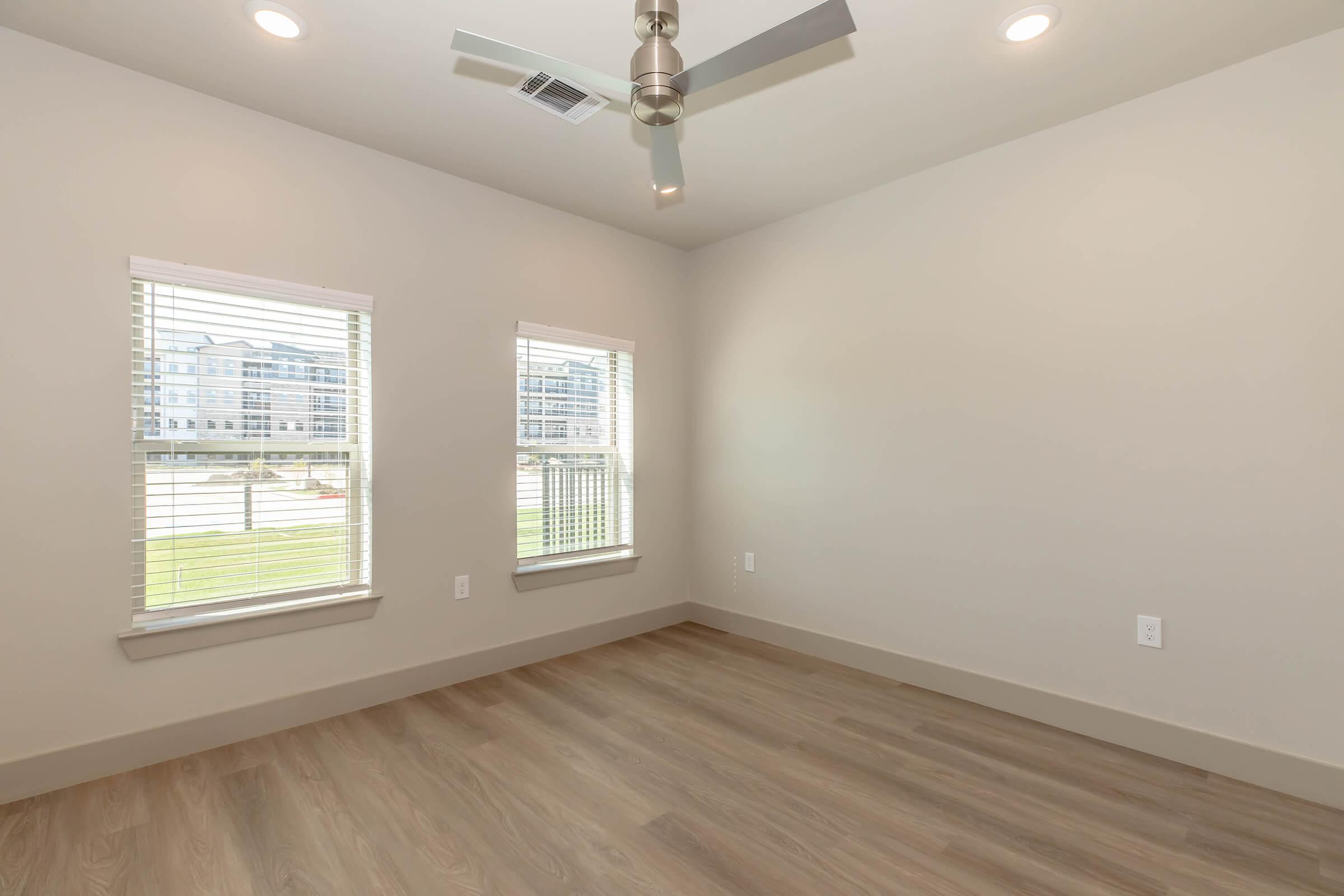 A bright, empty room with light gray walls and two windows featuring blinds. The room has a ceiling fan and light-colored laminate flooring. Natural light filters in from the windows, highlighting the spaciousness of the area.