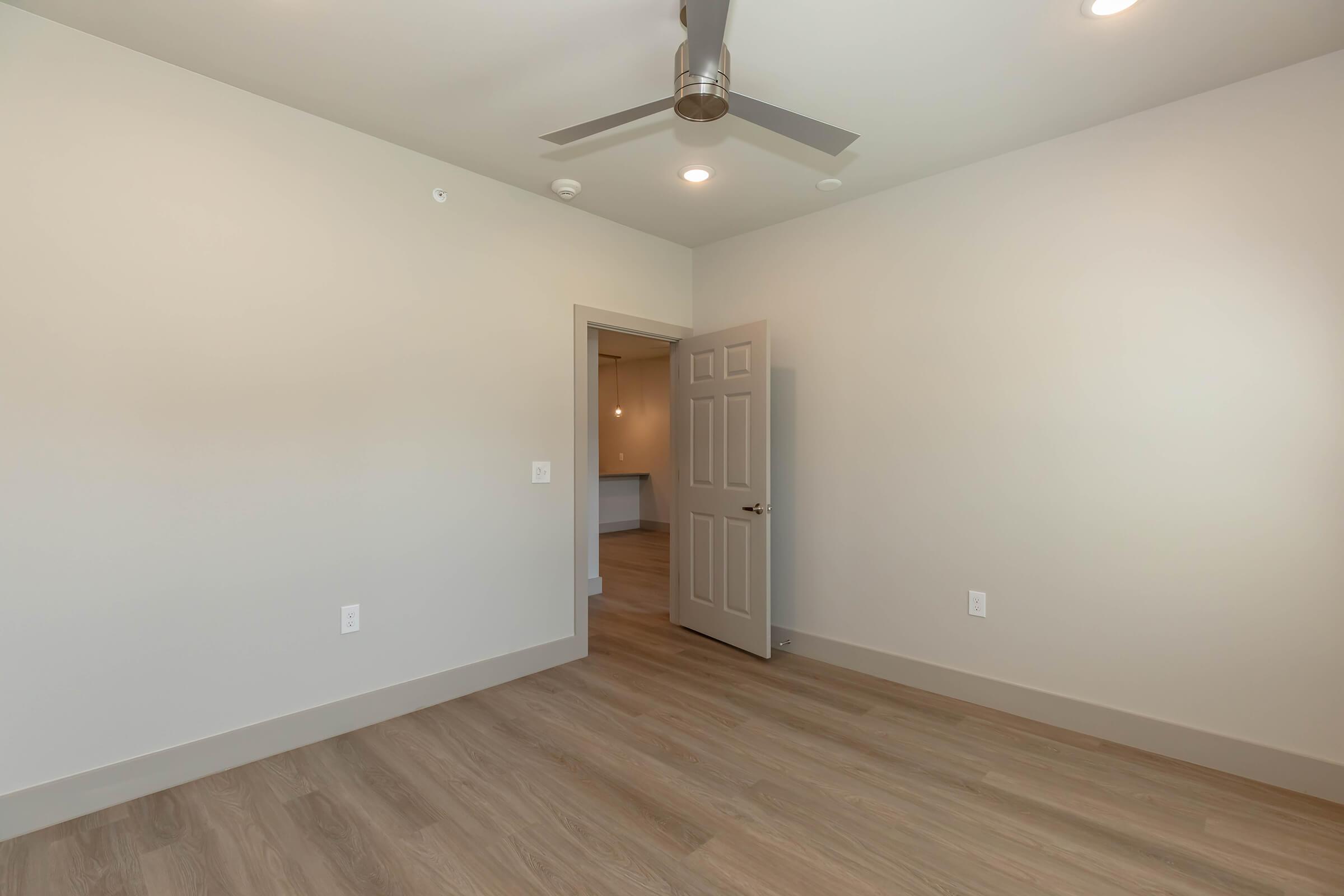 A spacious room with a light-colored wall and hardwood floor. There's a ceiling fan and a doorway leading to another area. The lighting is soft and ambient, creating an inviting atmosphere.