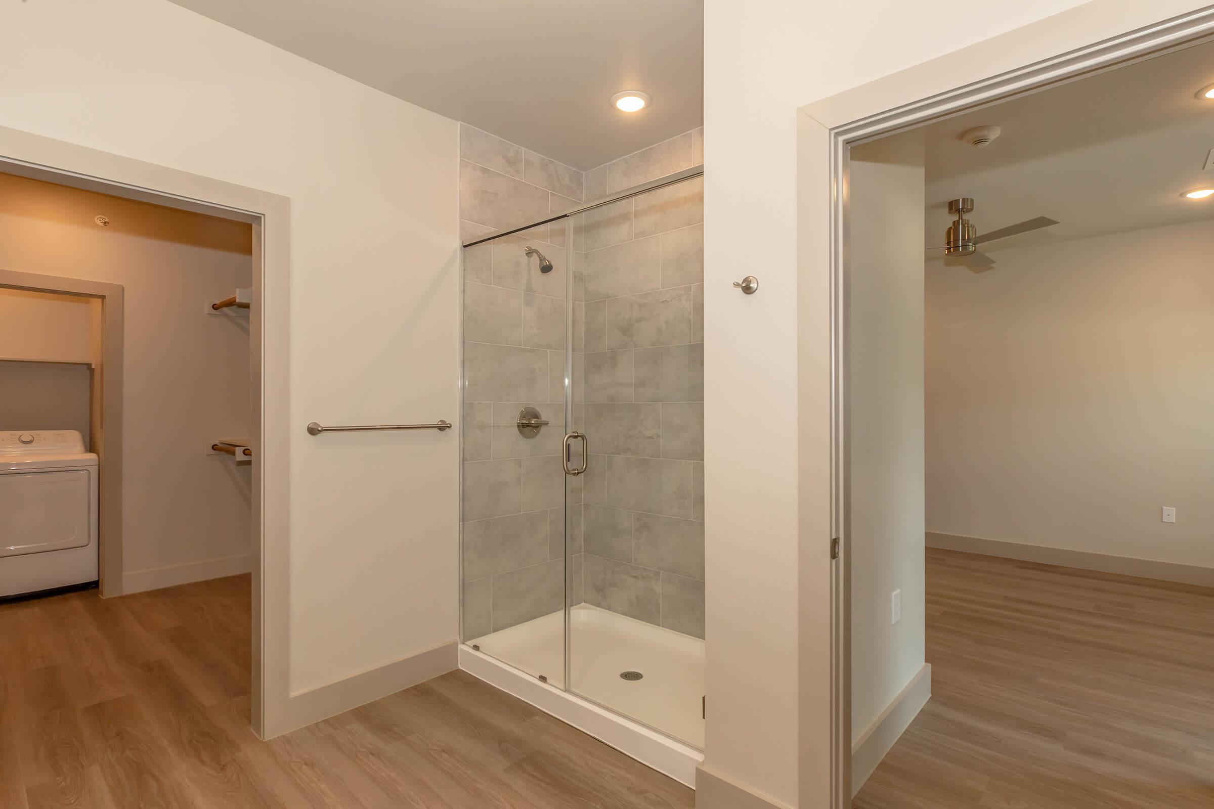 A modern bathroom featuring a walk-in shower with glass doors, light gray tile, and a sleek design. The view includes adjacent rooms, with a laundry area visible through an open door and a spacious, well-lit living area beyond. The flooring is a warm wood-like laminate.