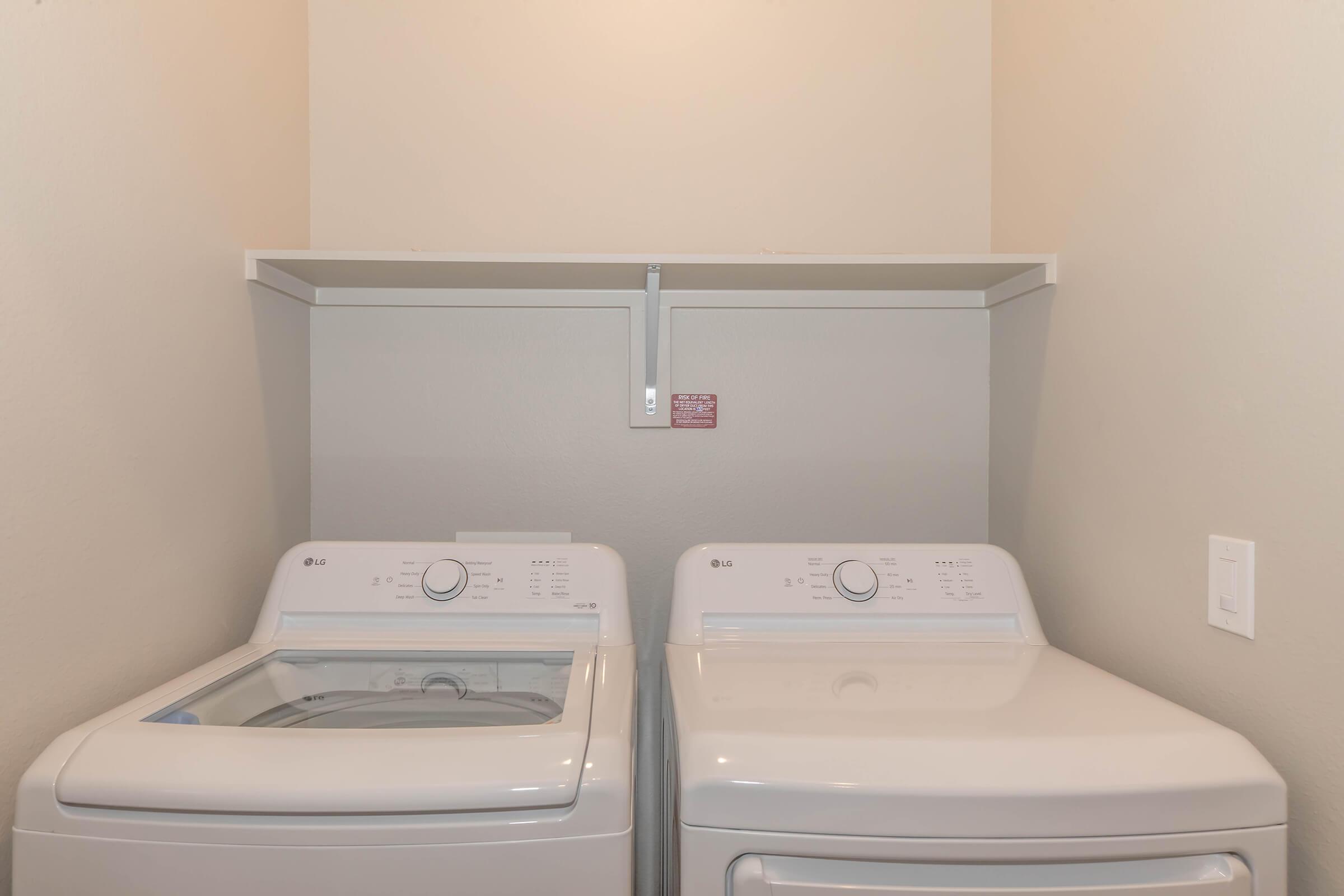 A clean laundry room featuring an LG washing machine on the left and an LG dryer on the right. Above them is a mounted shelf and a wall with a small warning sign. The walls are painted a light color, providing a bright and organized appearance.