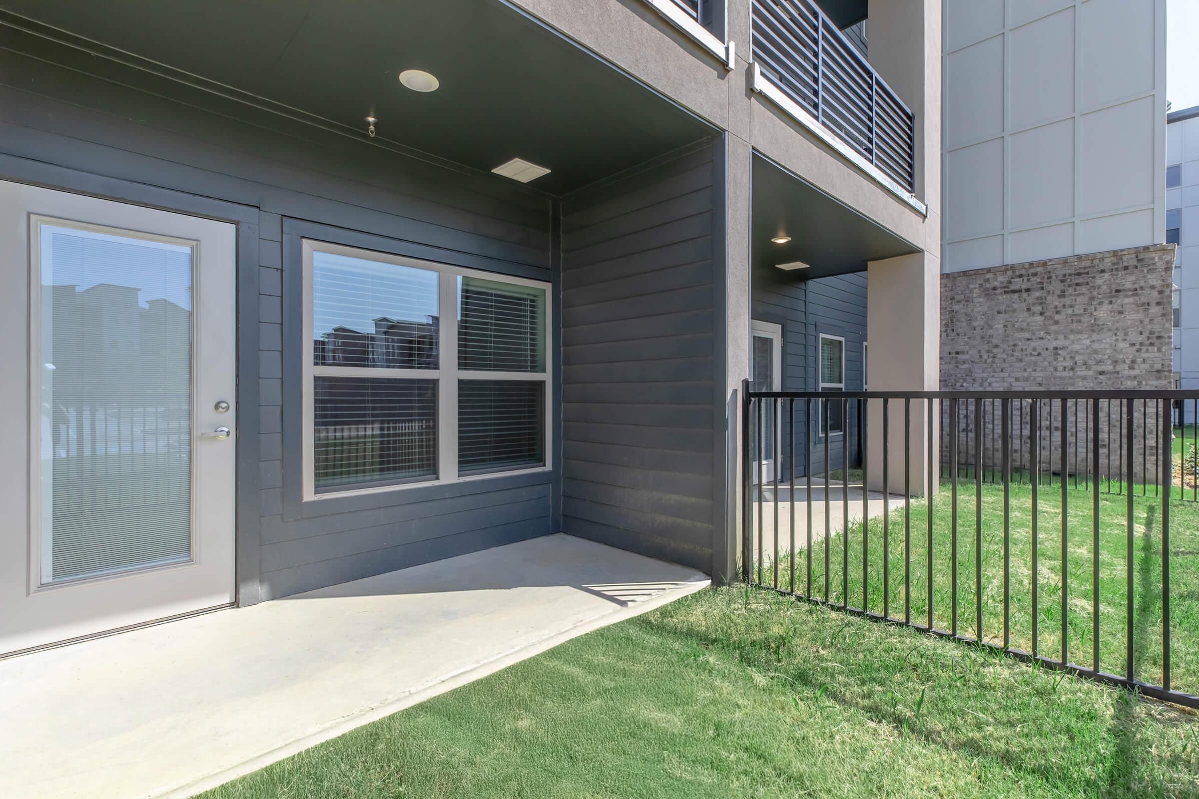 Exterior view of an apartment building featuring a patio area with grass, a fenced boundary, and large windows. The building has a modern design with a gray exterior, and there are two entrances visible. The setting looks clean and well-maintained, suitable for outdoor relaxation.