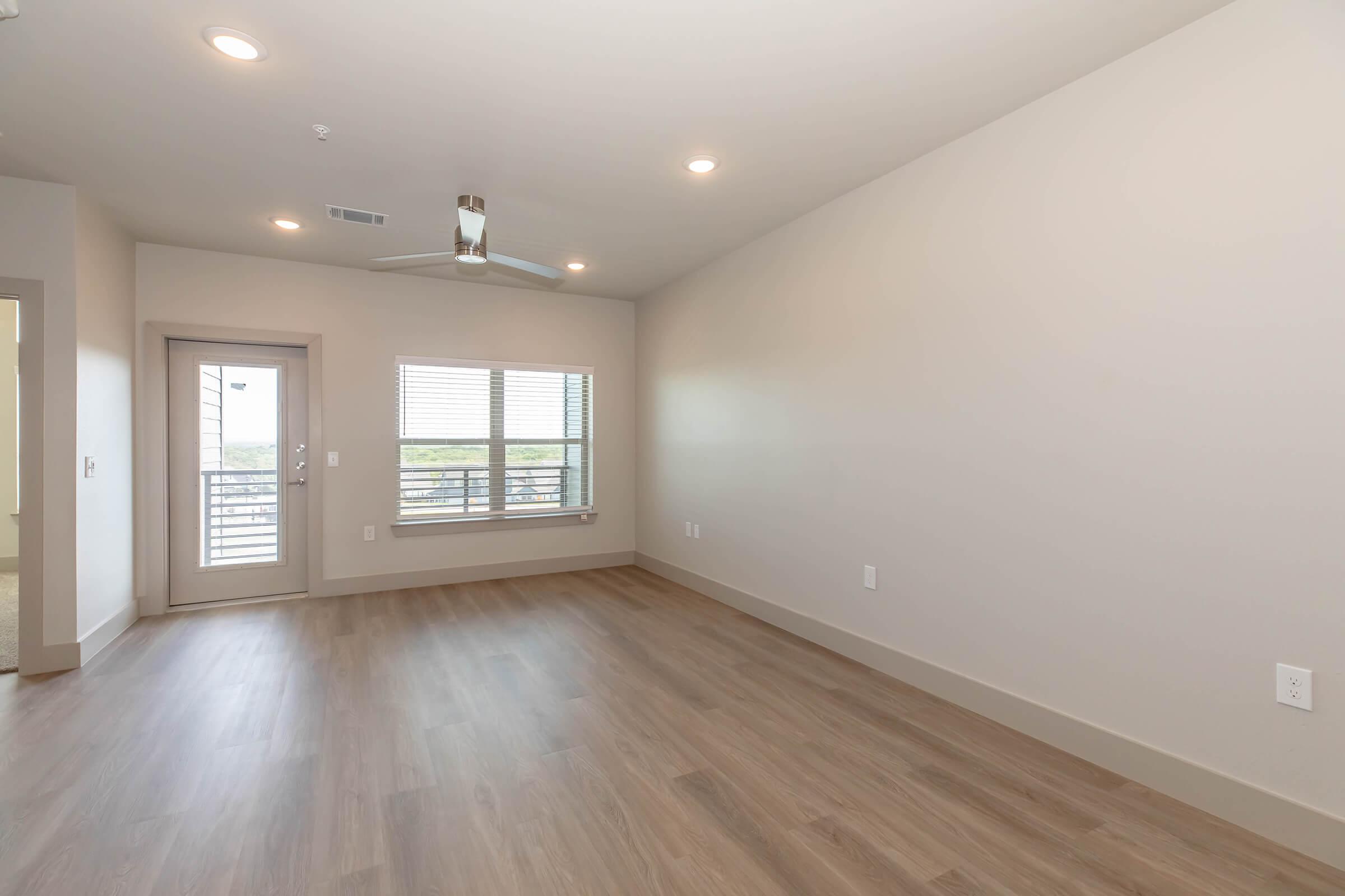 A spacious, empty living room with light wood flooring, neutral-colored walls, and a ceiling fan. A single set of glass sliding doors leads to a small balcony, and large windows allow natural light to fill the space. The room is well-lit with recessed ceiling lights, creating a modern and inviting atmosphere.
