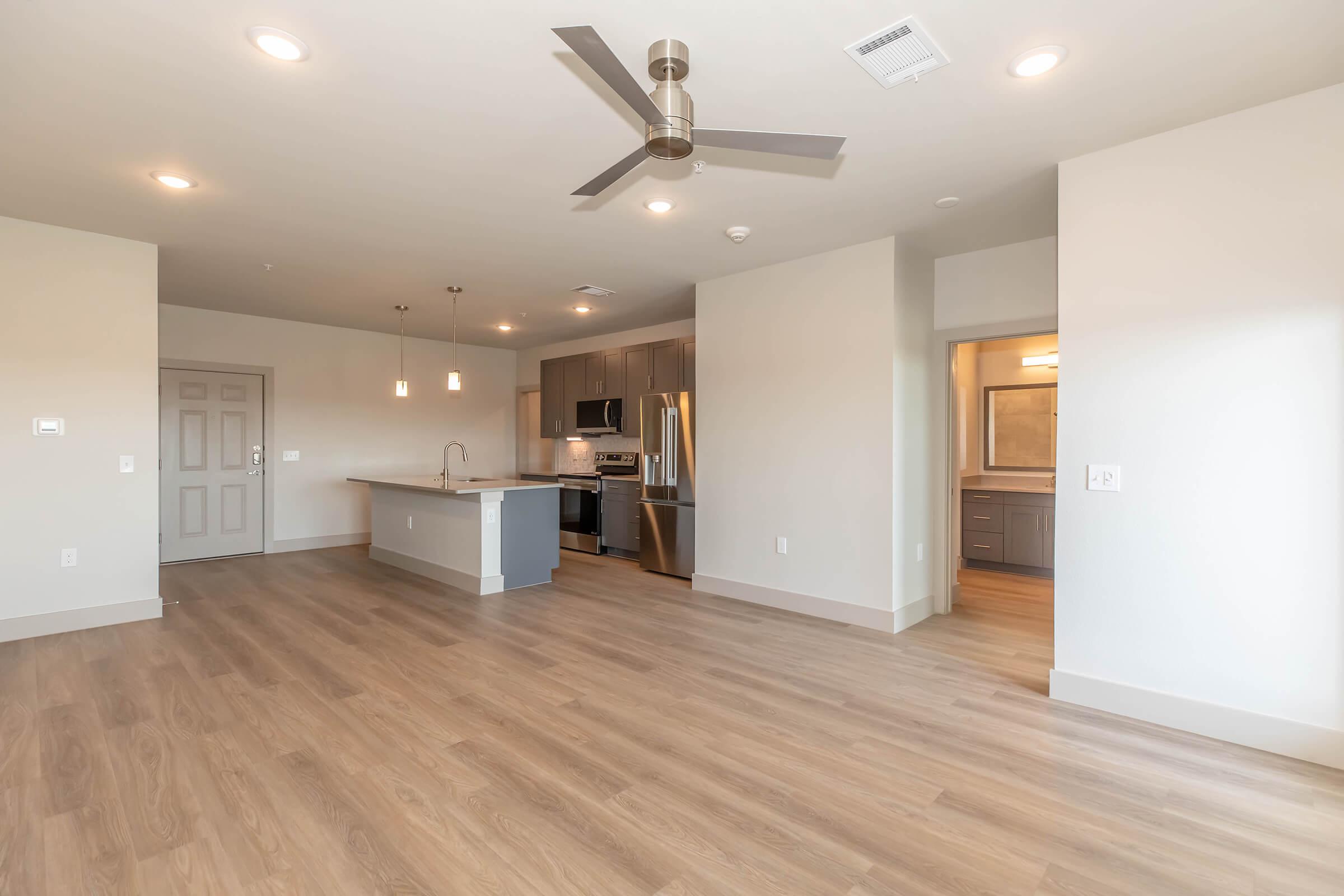 Modern open-concept apartment interior featuring a spacious living area with wooden flooring, a ceiling fan, and natural light. The kitchen includes stainless steel appliances and a central island, with a doorway leading to a bathroom. Neutral color palette enhances the contemporary design.