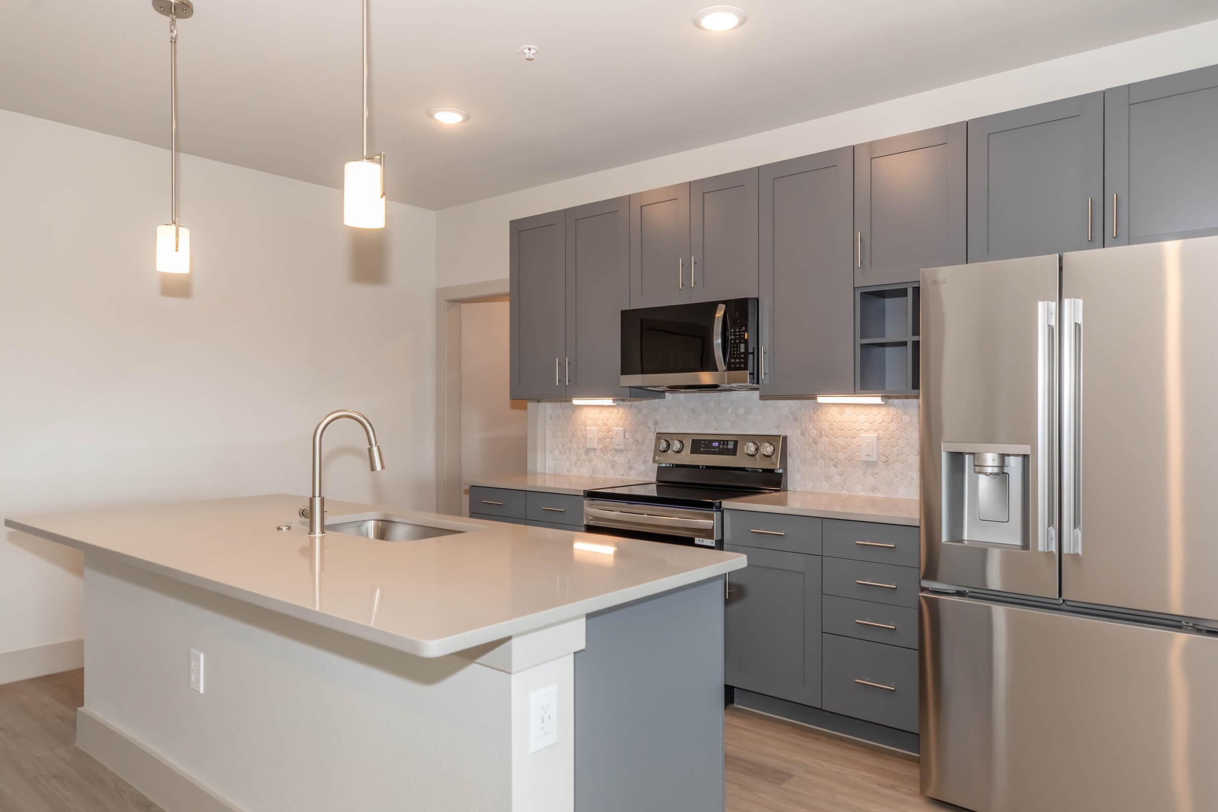Modern kitchen with gray cabinetry, stainless steel appliances, and a large island with a sink. Light-colored walls and warm wood flooring provide a contemporary atmosphere. Pendant lights hang above the island, and a tiled backsplash complements the sleek design.