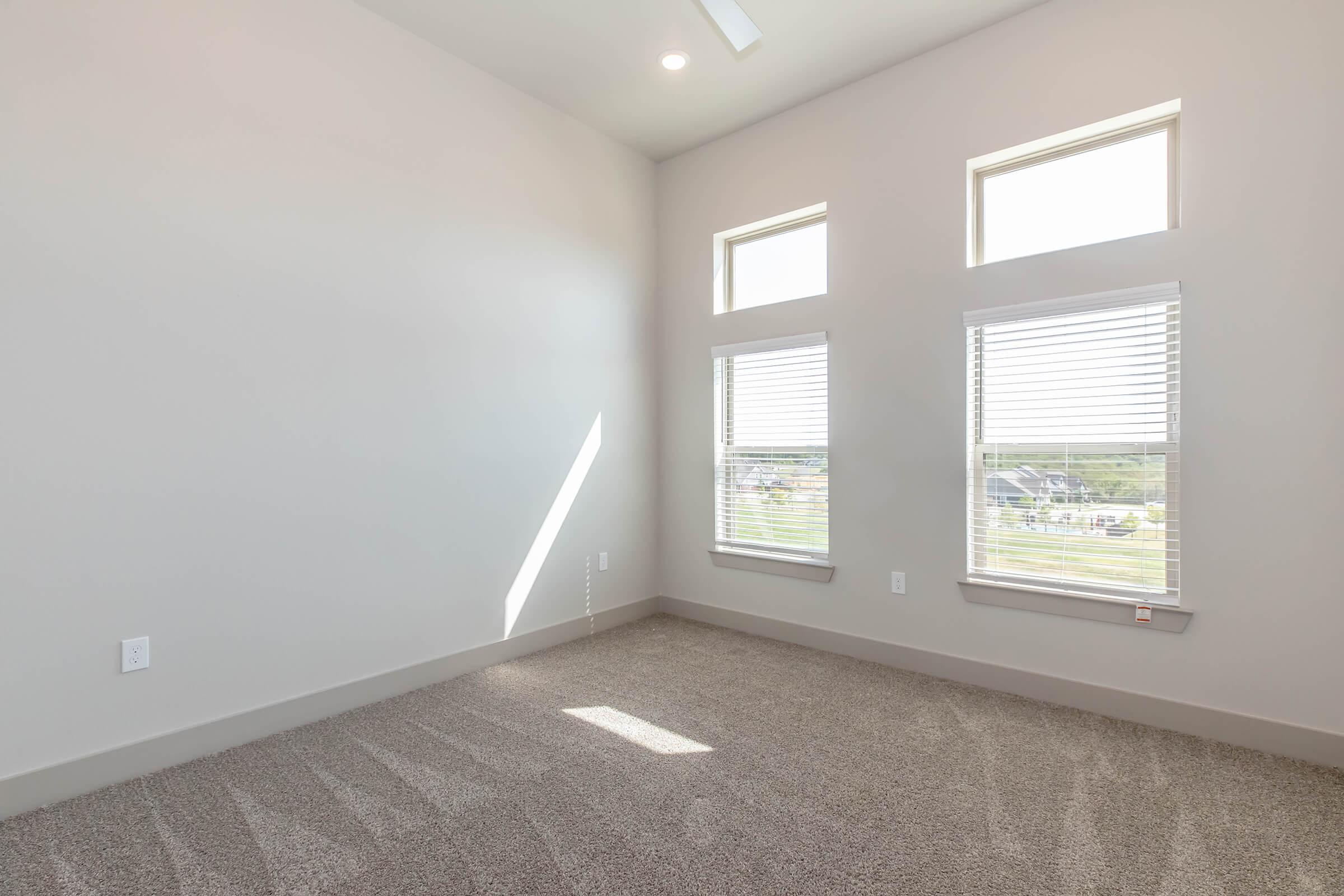 A bright, empty room with light gray walls and three windows with blinds. The floor is covered in light-colored carpet, and sunlight streams through the windows, creating a warm atmosphere. The room has a modern feel, with a light fixture on the ceiling and no furniture present.