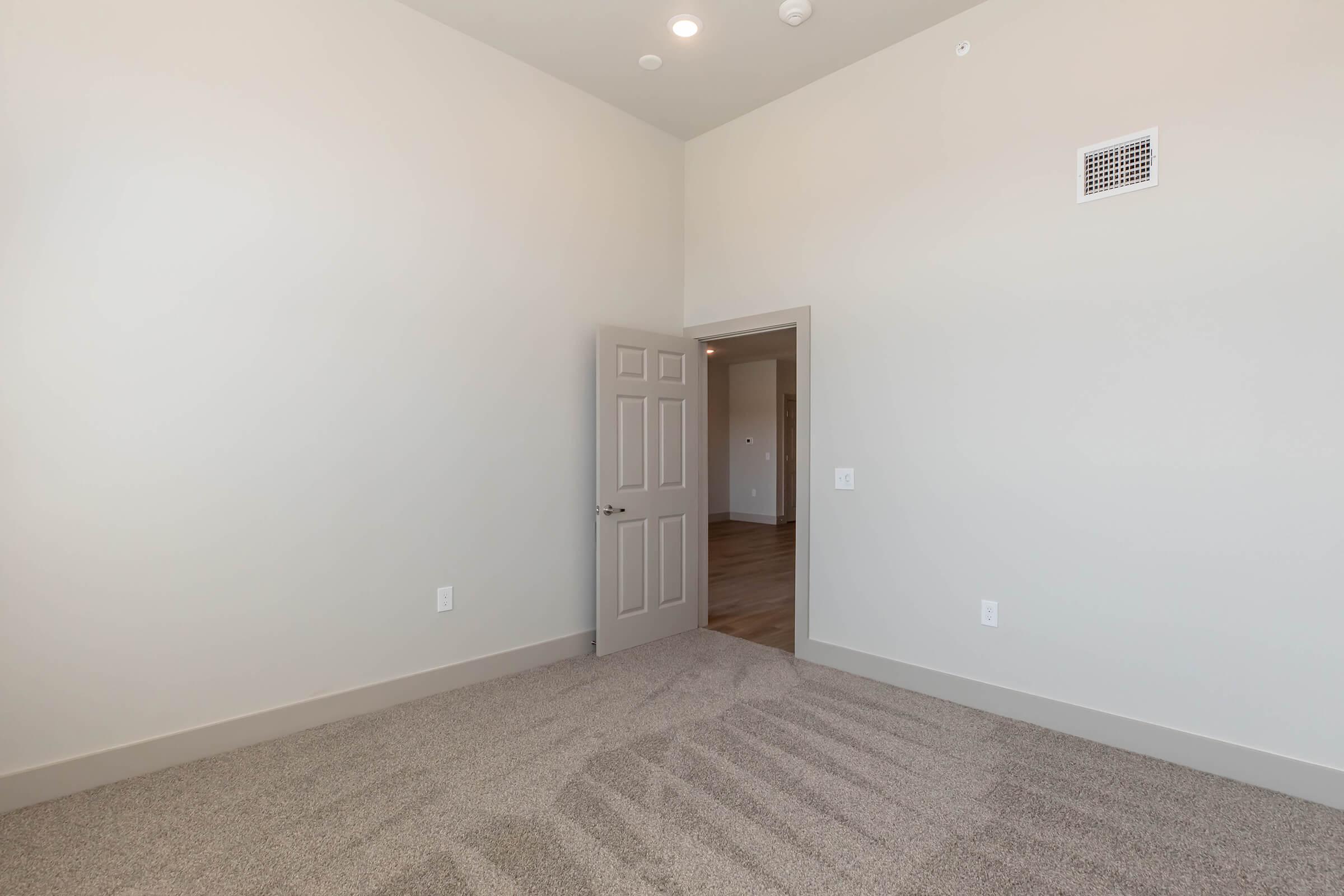 A spacious, empty room with light beige walls and gray carpeted flooring. A closed door on the left leads to another area, and the high ceiling features recessed lighting. The room appears bright and airy, showcasing a minimalist design suitable for various uses.
