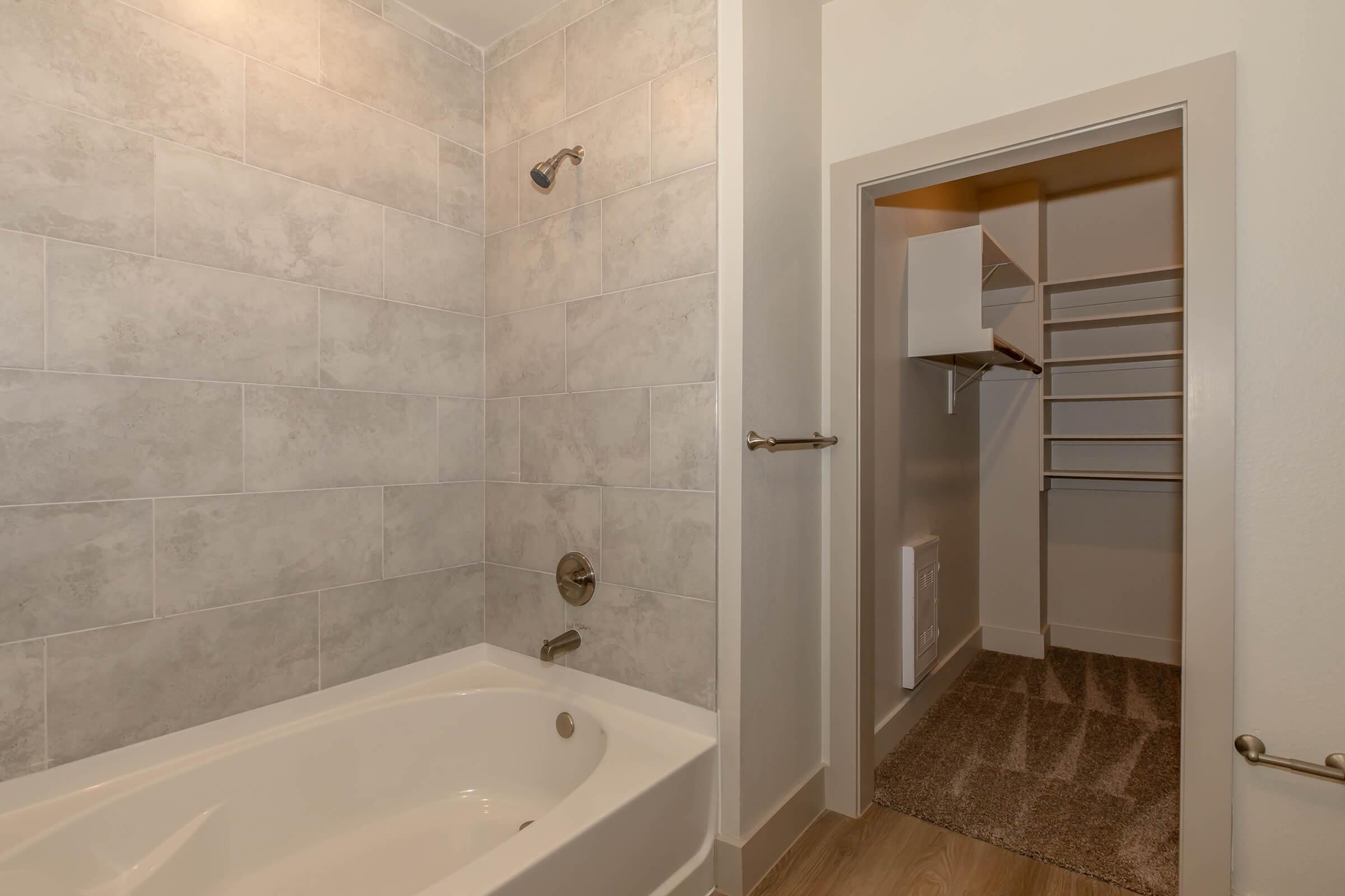 A modern bathroom featuring a bathtub with a shower head, gray tiled walls, and a light-colored floor. To the right, there's a walk-in closet with shelves and a carpeted floor, creating a spacious and organized feel. The overall design is clean and contemporary.