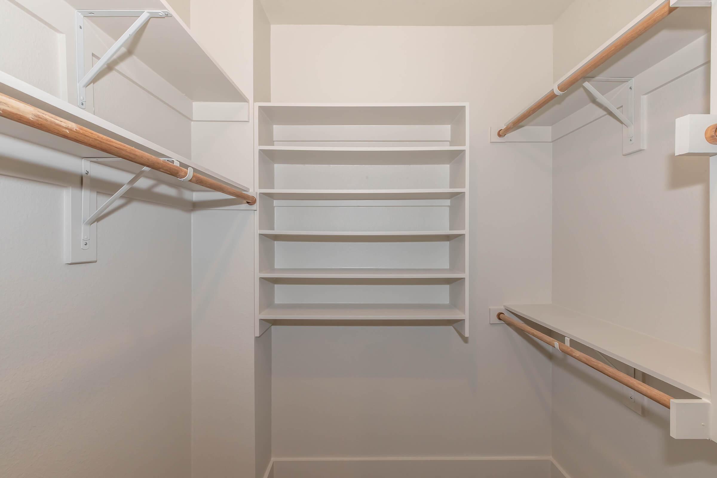 A spacious empty closet featuring two hanging rods and multiple shelving units against a white wall. The shelving is arranged to maximize storage space, providing a clean and organized look. The floor is clear, emphasizing the room's ample space for storage or organization.