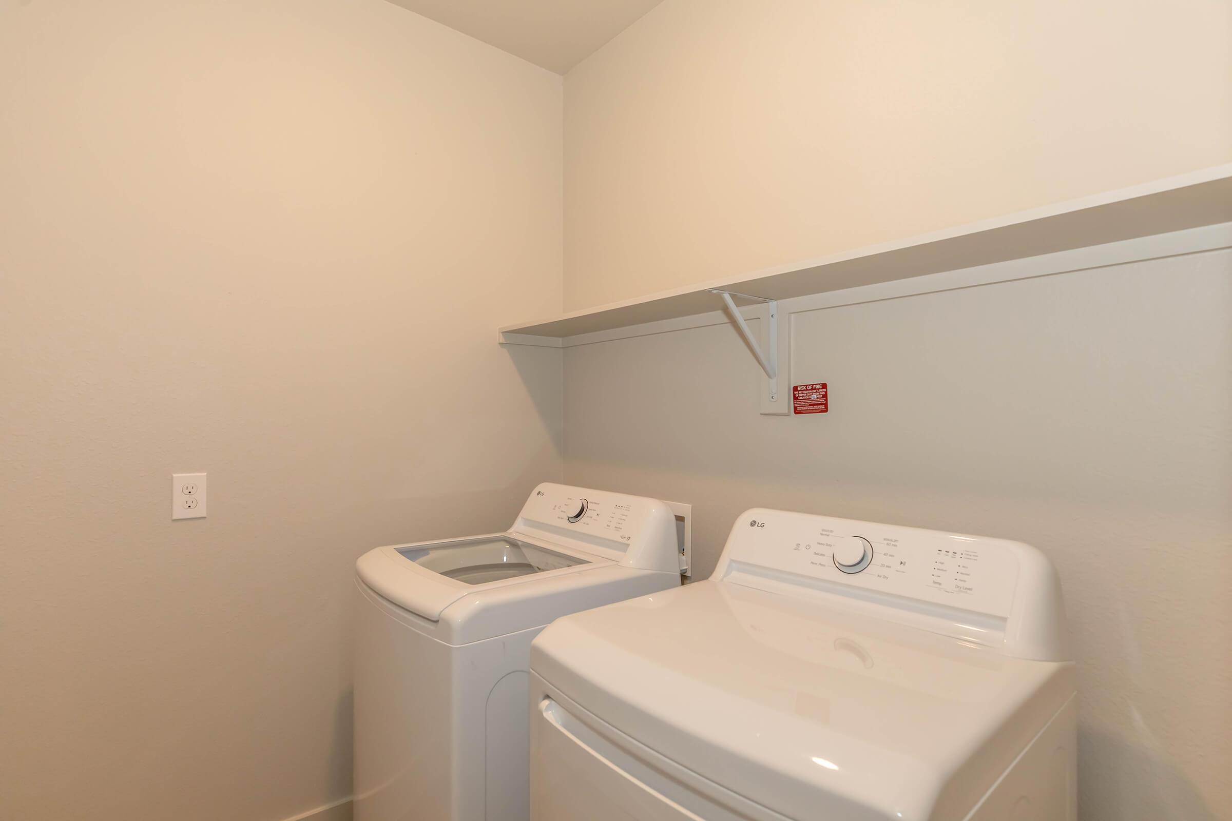 A laundry room featuring two white washing machines side by side, with a minimalistic design. Above the machines, there is a white shelf and a red warning sign on the wall. The walls are painted a light color, contributing to a clean and tidy atmosphere.