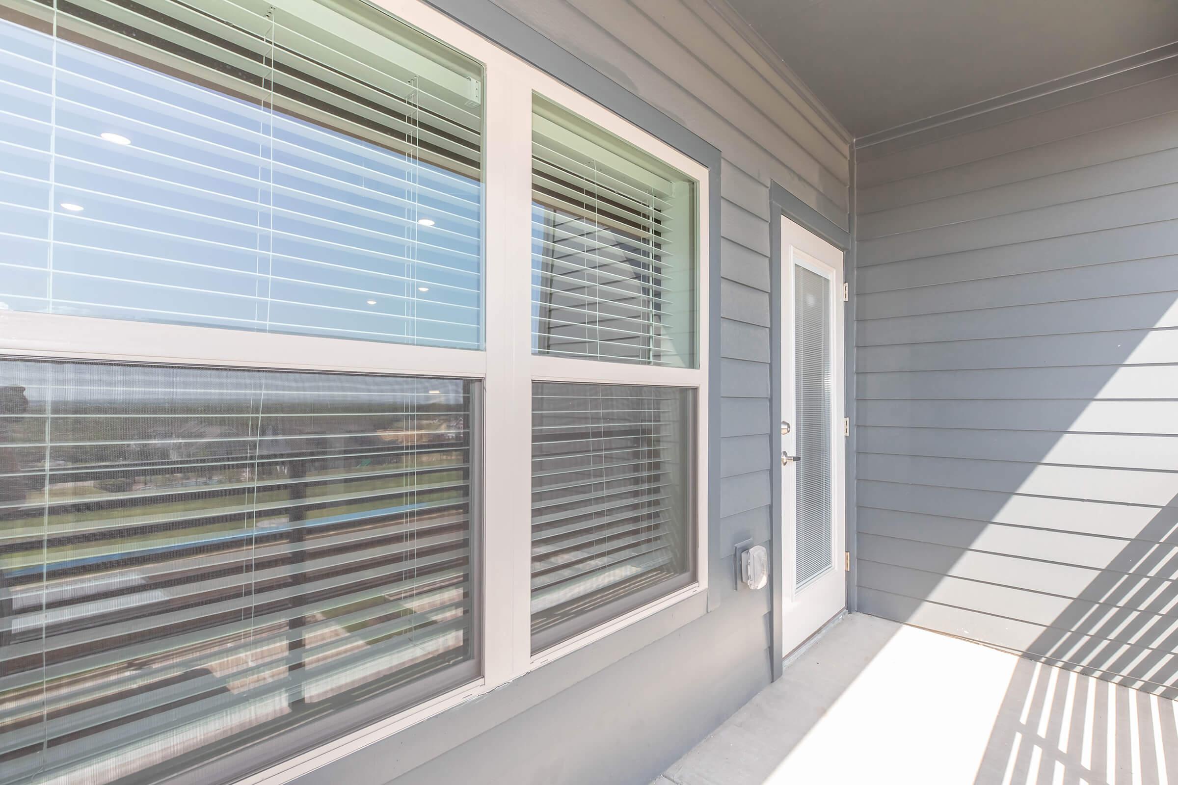 A corner view of a balcony featuring a wall with gray siding, large windows with white blinds, and a door that leads outside. Sunlight creates shadows along the surface, highlighting the clean lines and modern design of the space.