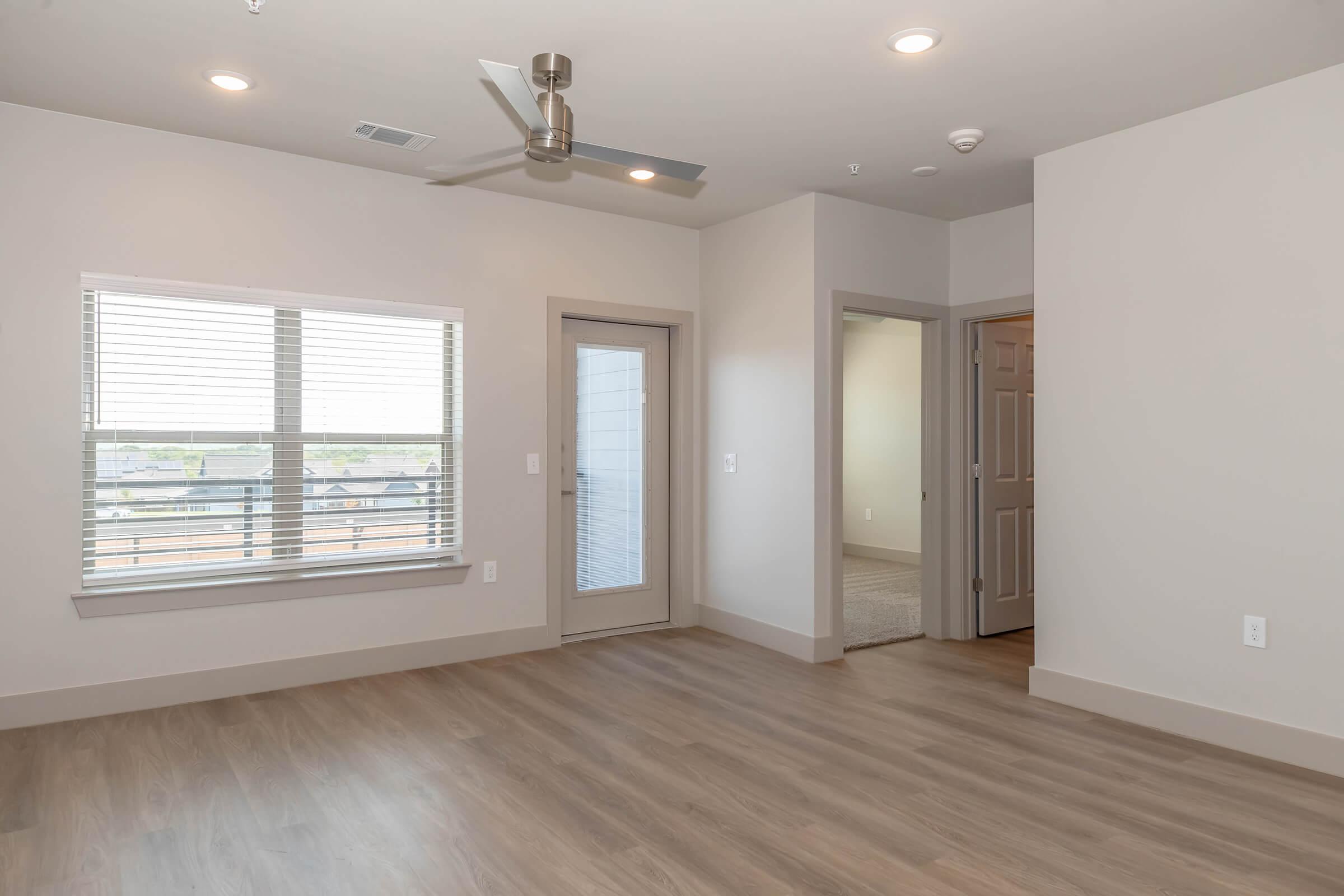 A well-lit, modern interior of a spacious room featuring light-colored walls, a ceiling fan, and large windows with blinds. There is a door leading outside and a doorway to another room, with hardwood-style flooring throughout. The space appears empty and clean, suitable for various arrangements.