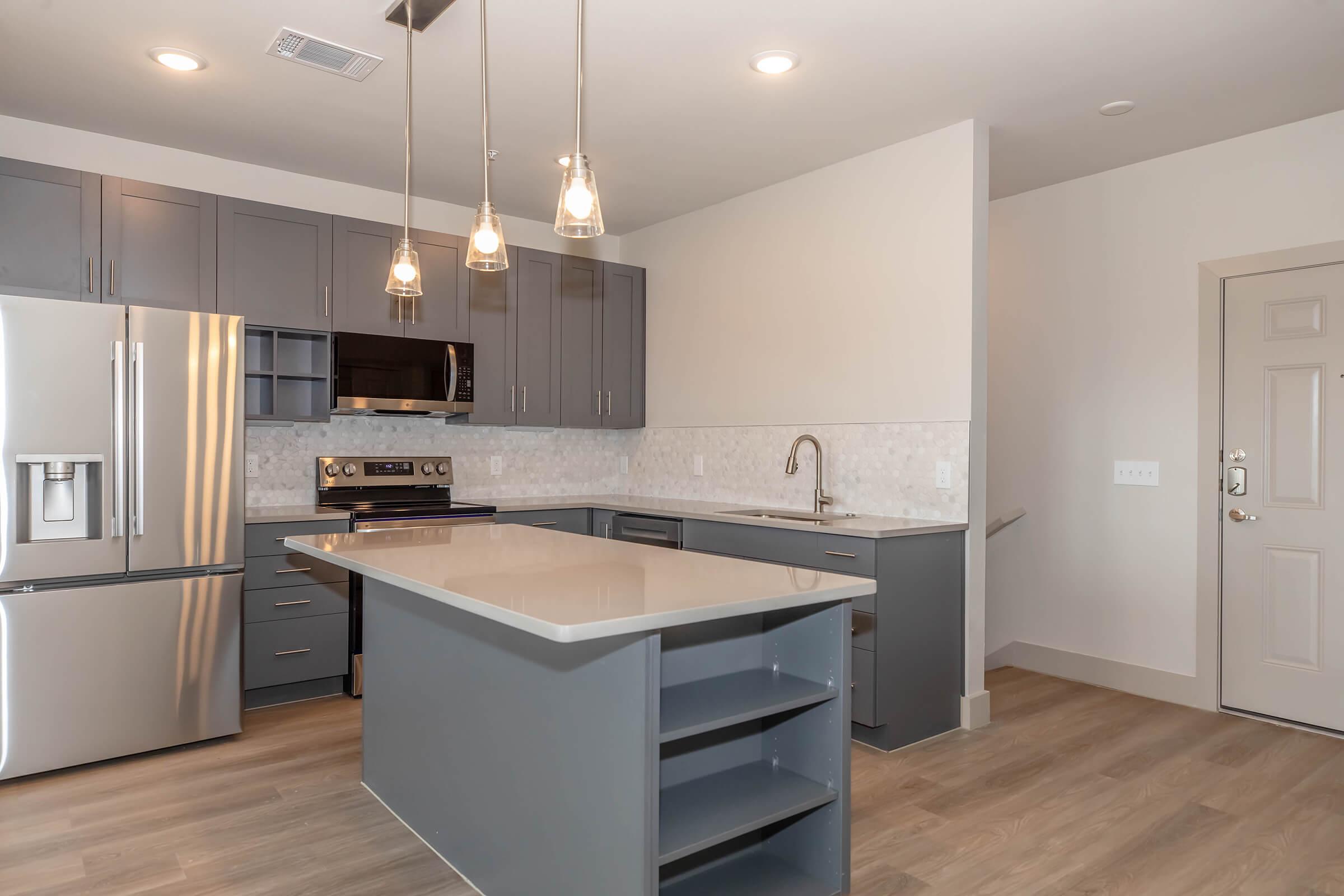 Modern kitchen featuring gray cabinetry, stainless steel appliances, an island with seating, and pendant lighting. The walls are a neutral tone, and there is a door leading to another room. The flooring is light wood, contributing to a spacious and contemporary feel.