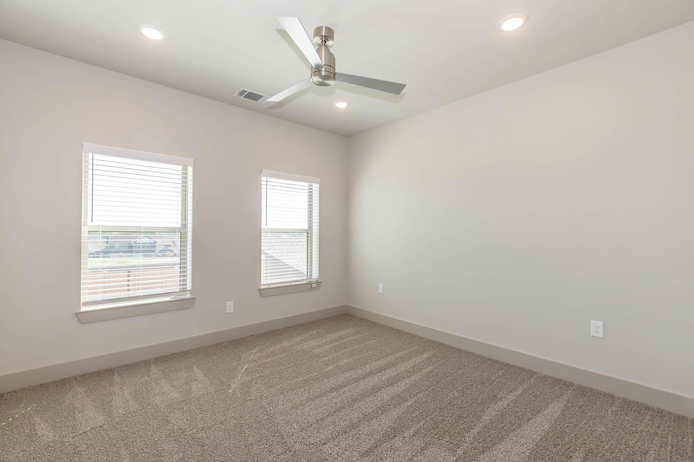A bright, empty room featuring light gray walls, two windows with white blinds, a ceiling fan, and carpeted flooring. The space has a modern feel with natural light streaming in, creating a clean and open atmosphere.