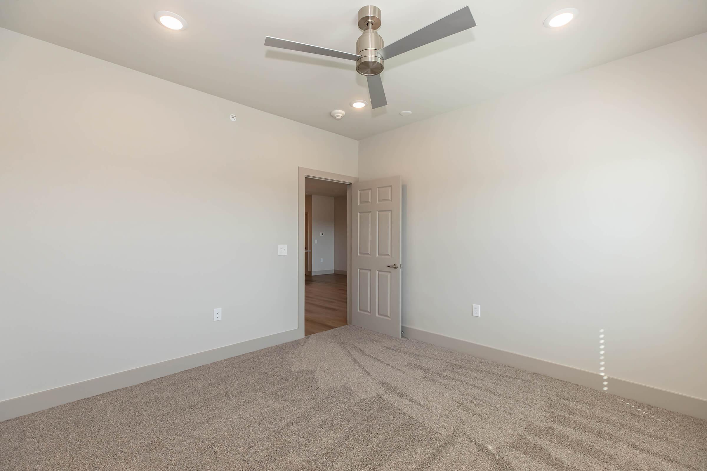 A spacious, empty room with light beige walls and a ceiling fan. The floor is covered with light-colored carpet. There's a door leading to another room on the left side, and natural light enters through a window, creating a bright atmosphere.