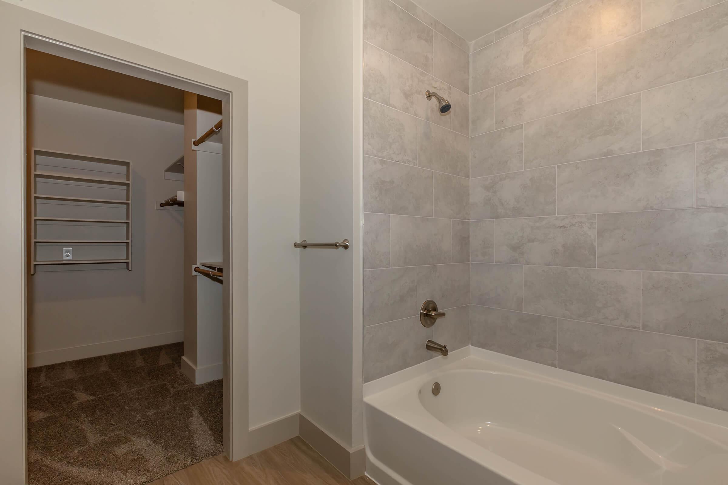 A modern bathroom featuring a bathtub with a showerhead, tiled walls in light gray tones, and a door leading to a closet area. The floor is covered with textured tiles, and the space has neutral-colored walls and fixtures, creating a clean and contemporary aesthetic.