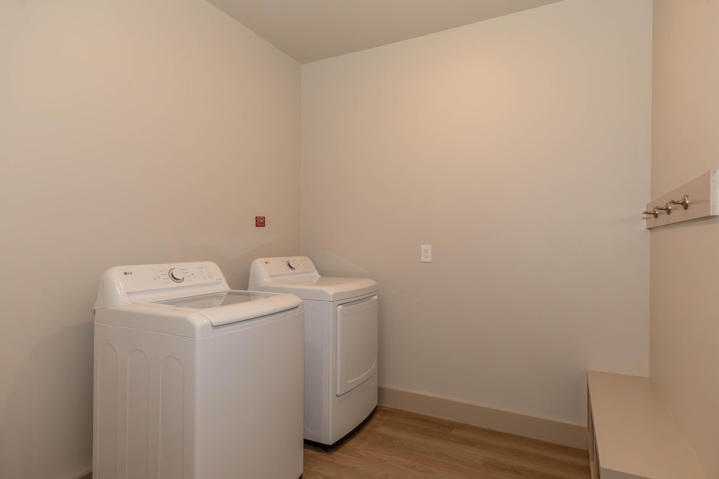 A laundry room featuring two white washing machines side by side, set against a light-colored wall. There is a small shelf with hooks on the wall and a light-colored floor. The space is bright and uncluttered, emphasizing a clean and modern design.