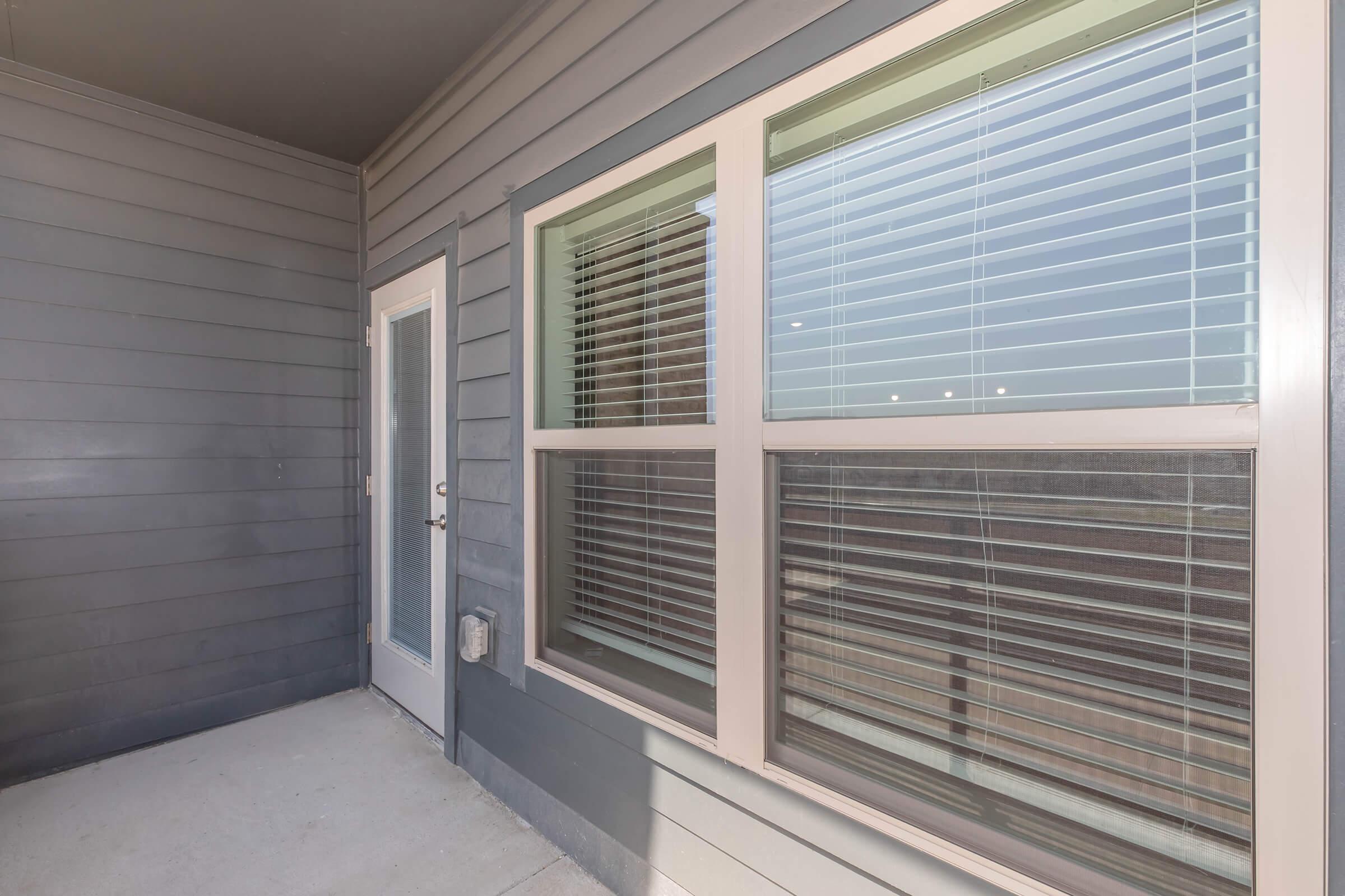 Exterior view of a modern balcony with gray siding, featuring a glass door and large windows with blinds. The concrete floor is visible, and the overall lighting suggests a clear day.