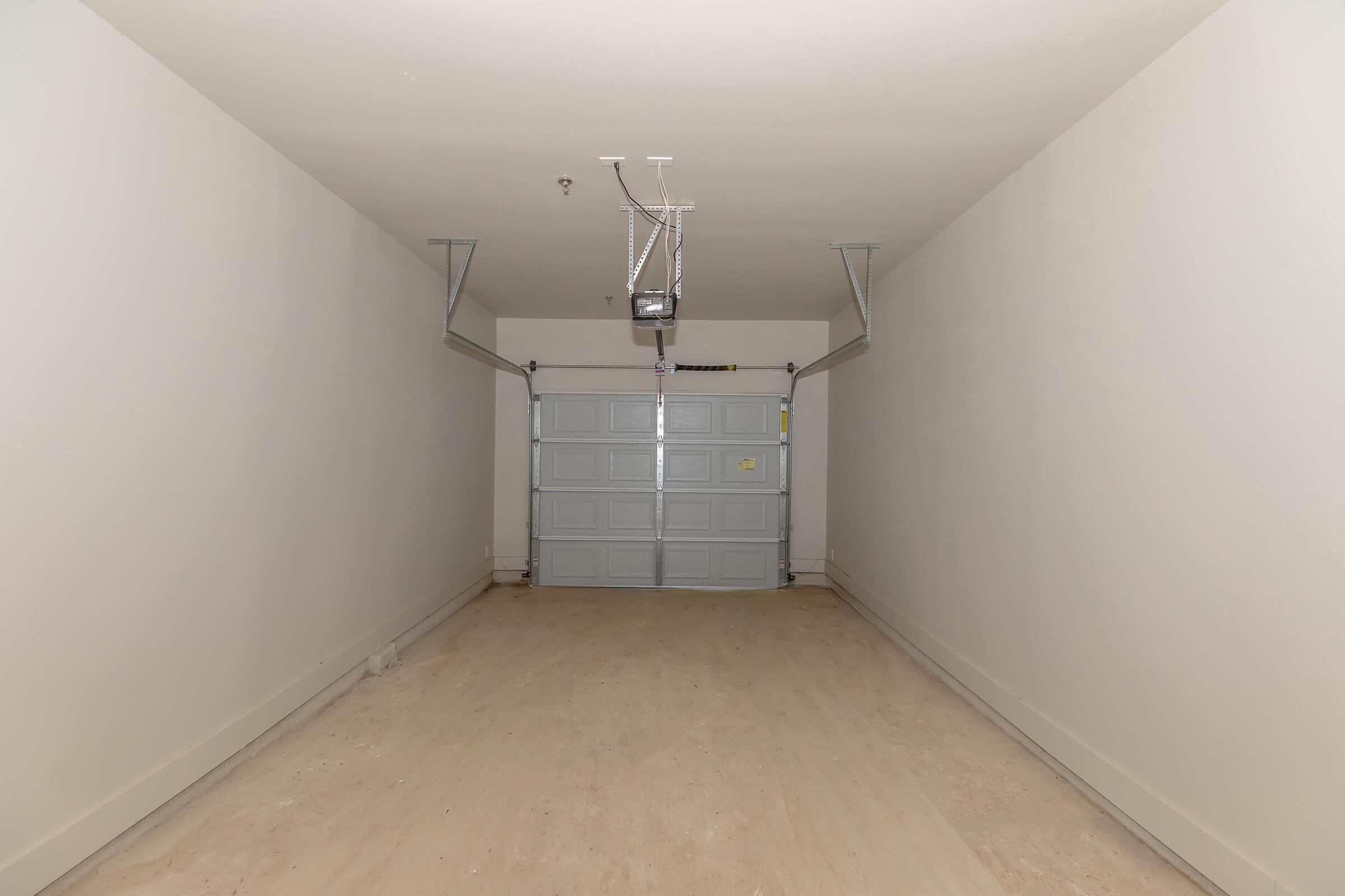 Interior of a spacious, empty garage featuring light-colored walls and a wooden floor. The garage door is closed and painted in a light blue color, with a ceiling-mounted garage door opener visible above. The space is well-lit, creating a clean and open atmosphere.