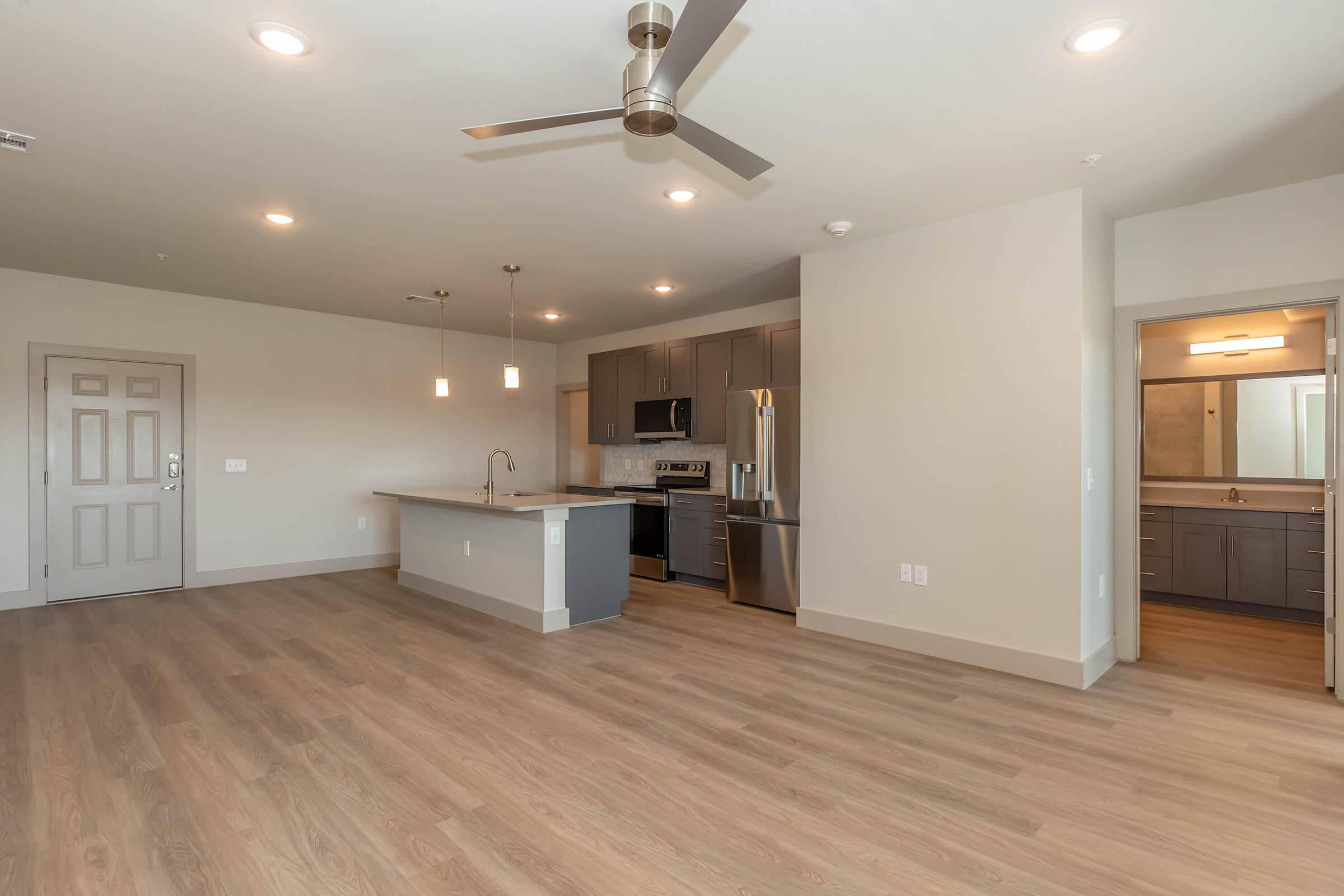 Modern open-concept living area featuring a kitchen with stainless steel appliances, a central island with seating, and a spacious hardwood floor. The room has recessed lighting, a ceiling fan, and is connected to a bathroom and hallway. Neutral color palette with ample natural light.