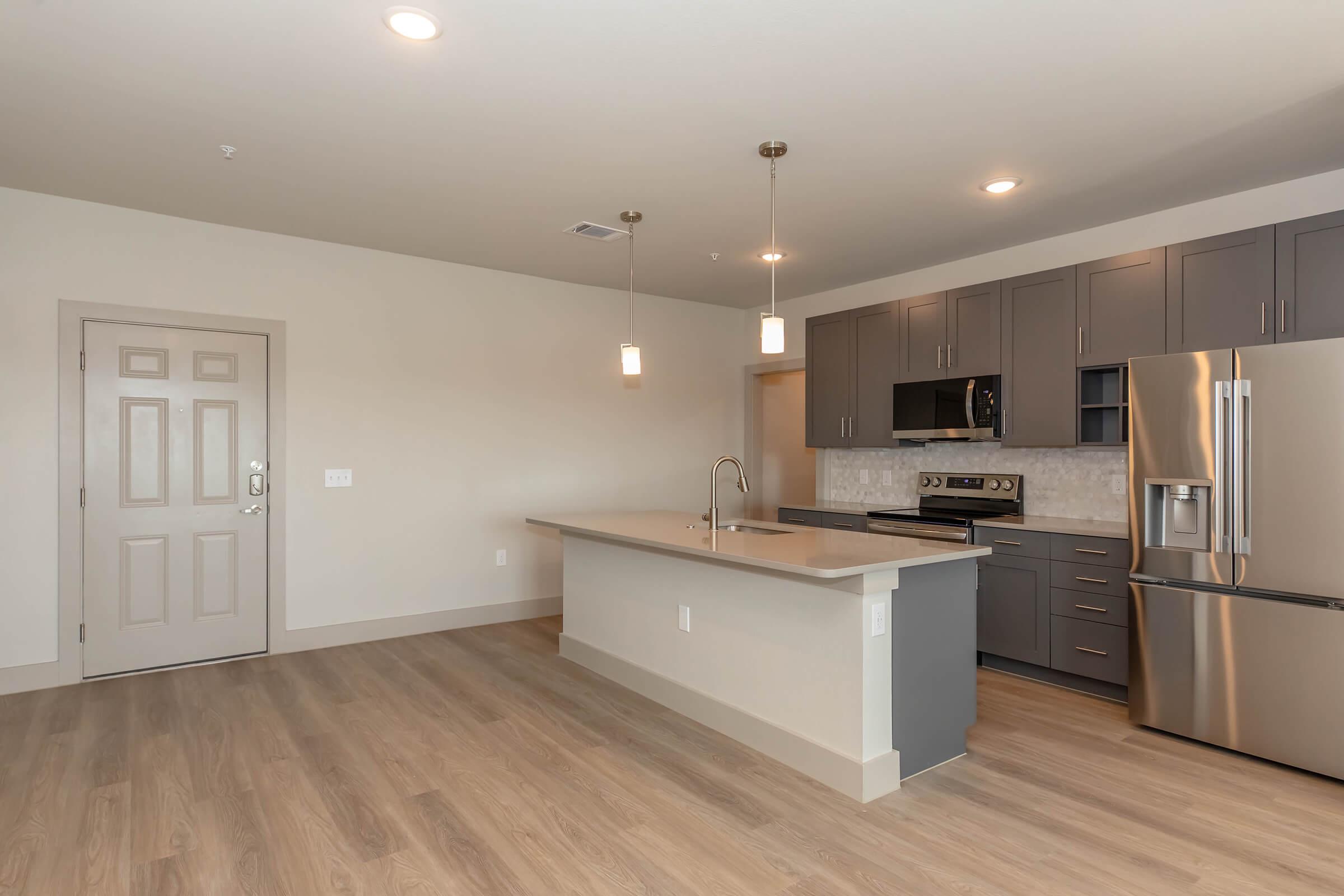 Modern kitchen in an open-concept layout, featuring grey cabinets, stainless steel appliances, and a large island with a sink. The space has warm wood-like flooring and pendant lighting above the island. A door leads to an adjacent room, offering a spacious and contemporary feel.