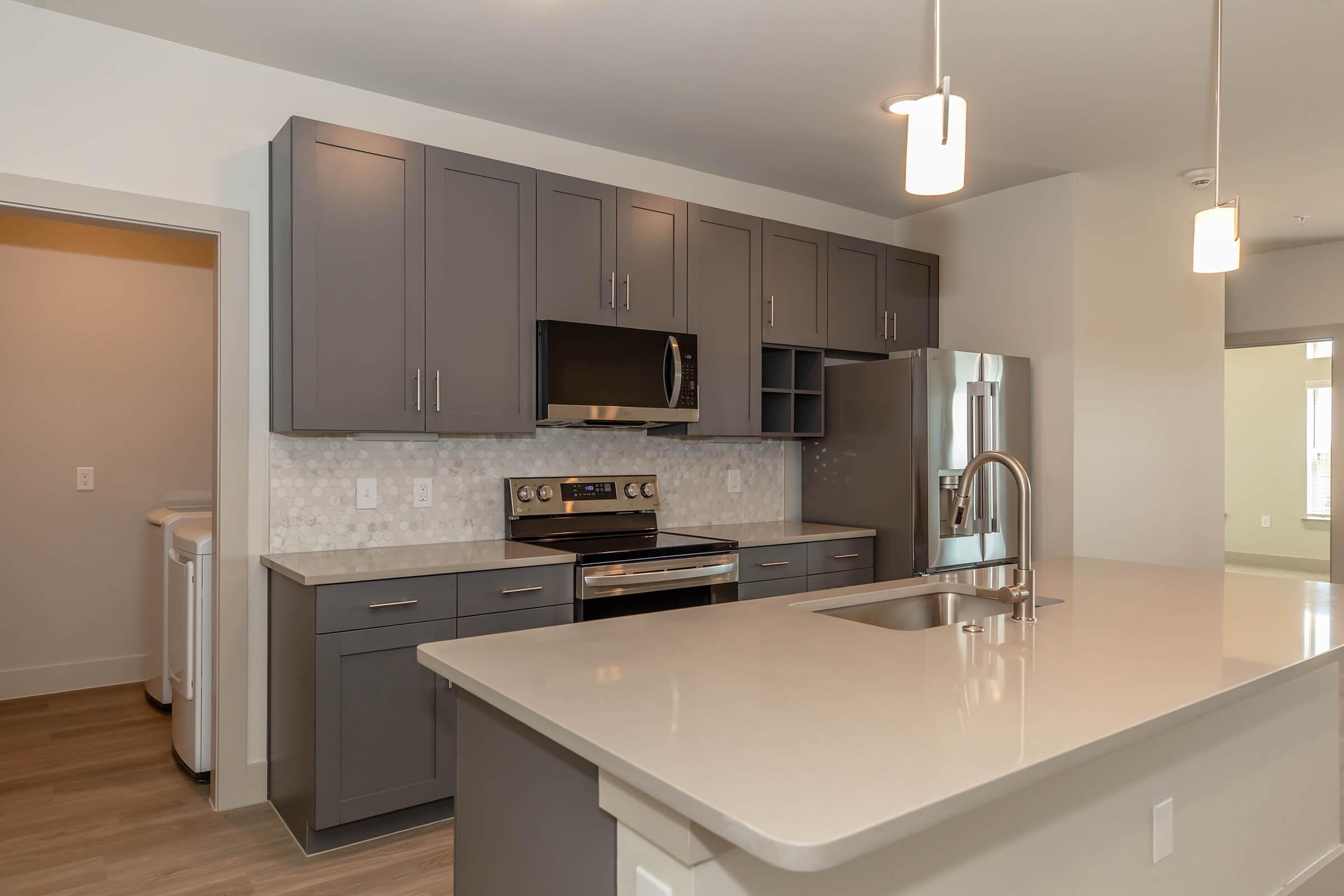 Modern kitchen featuring gray cabinets, stainless steel appliances, a microwave, and a large island with a sink. The backsplash is a light-colored mosaic. Soft lighting fixtures hang above the island, and there's a door leading to another room on the left. The overall aesthetic is clean and contemporary.