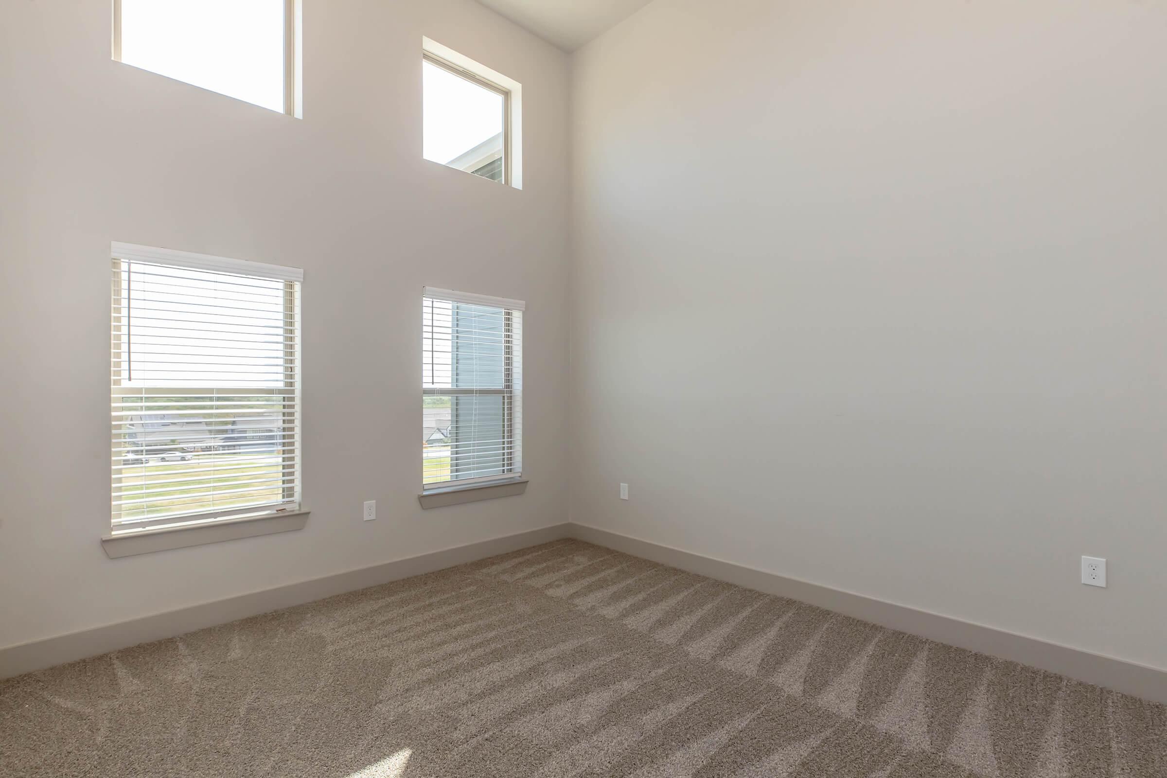 Empty room with light-colored walls and a beige carpet. There are four windows allowing natural light, with blinds partially closed. The room has a high ceiling and a simple, clean design, creating a spacious feel.