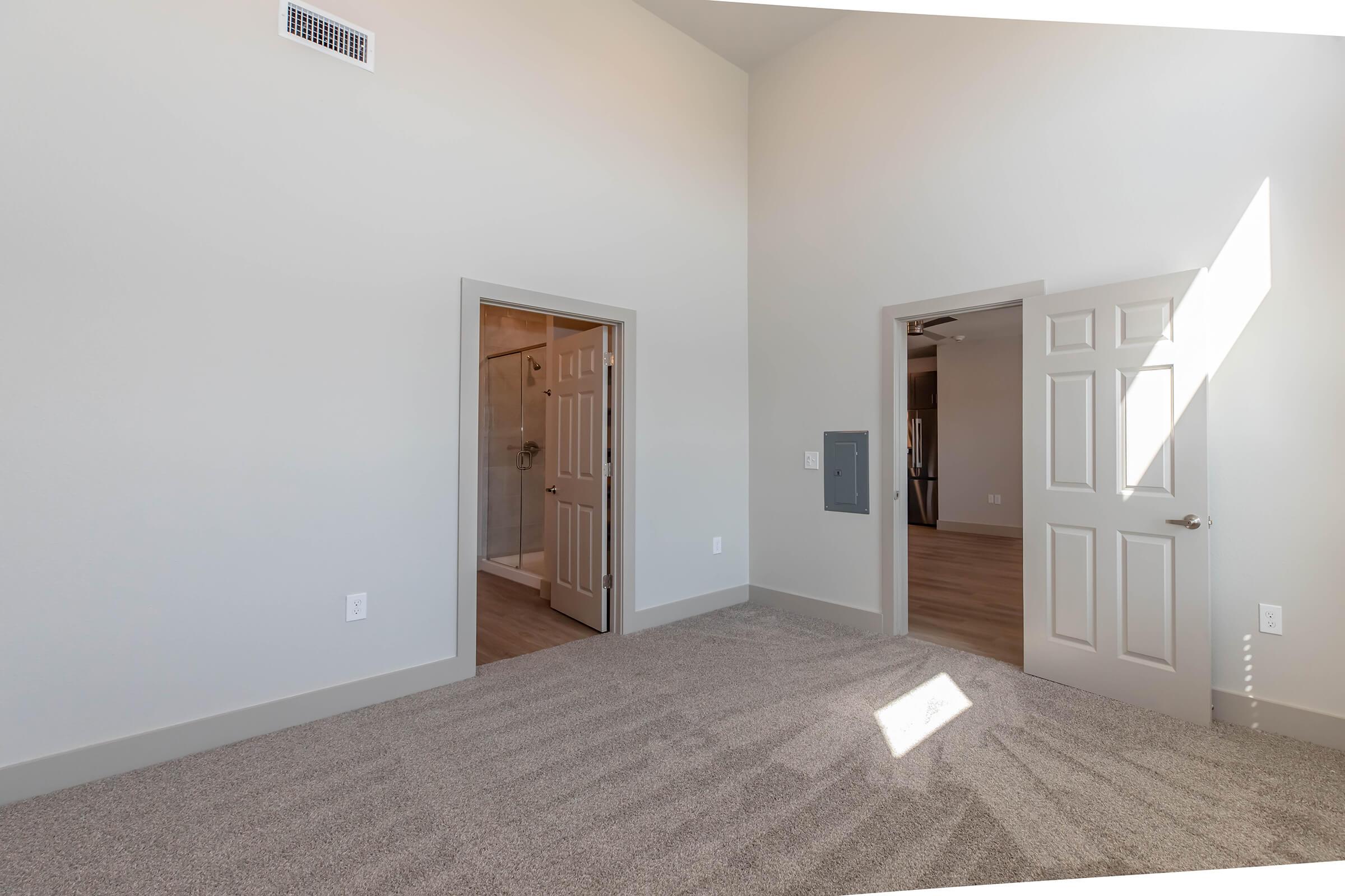 A spacious, well-lit room featuring neutral-colored walls and plush carpet. Two closed doors lead to additional spaces, while a large window allows natural light to fill the room. The ceiling is high, adding to the open feel of the area.