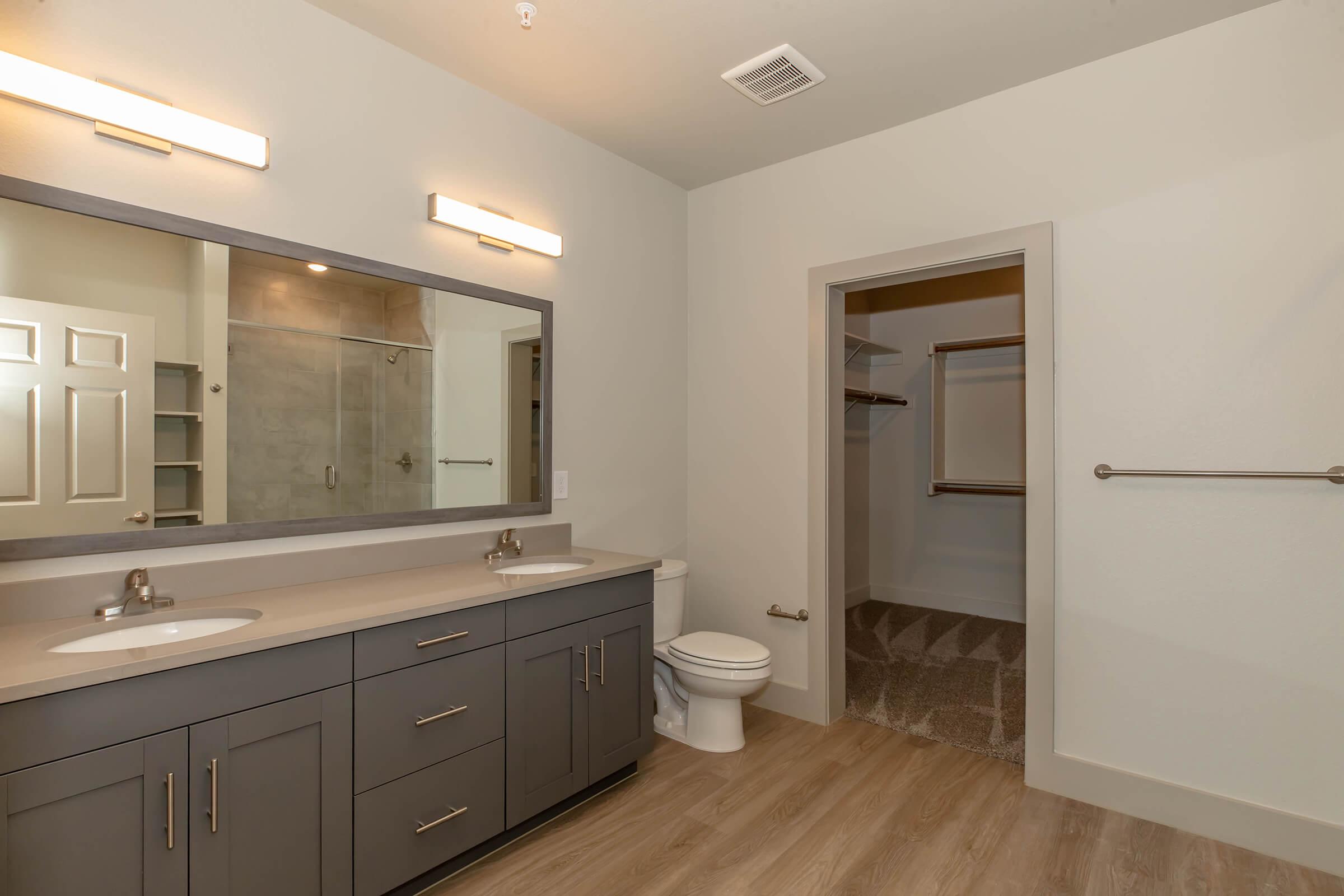 Modern bathroom featuring dual sinks with a large mirror, stylish lighting fixtures, a toilet, and a spacious shower area. The decor includes a neutral color palette and wooden flooring, with an adjacent walk-in closet visible. Overall, it presents a clean and contemporary design.