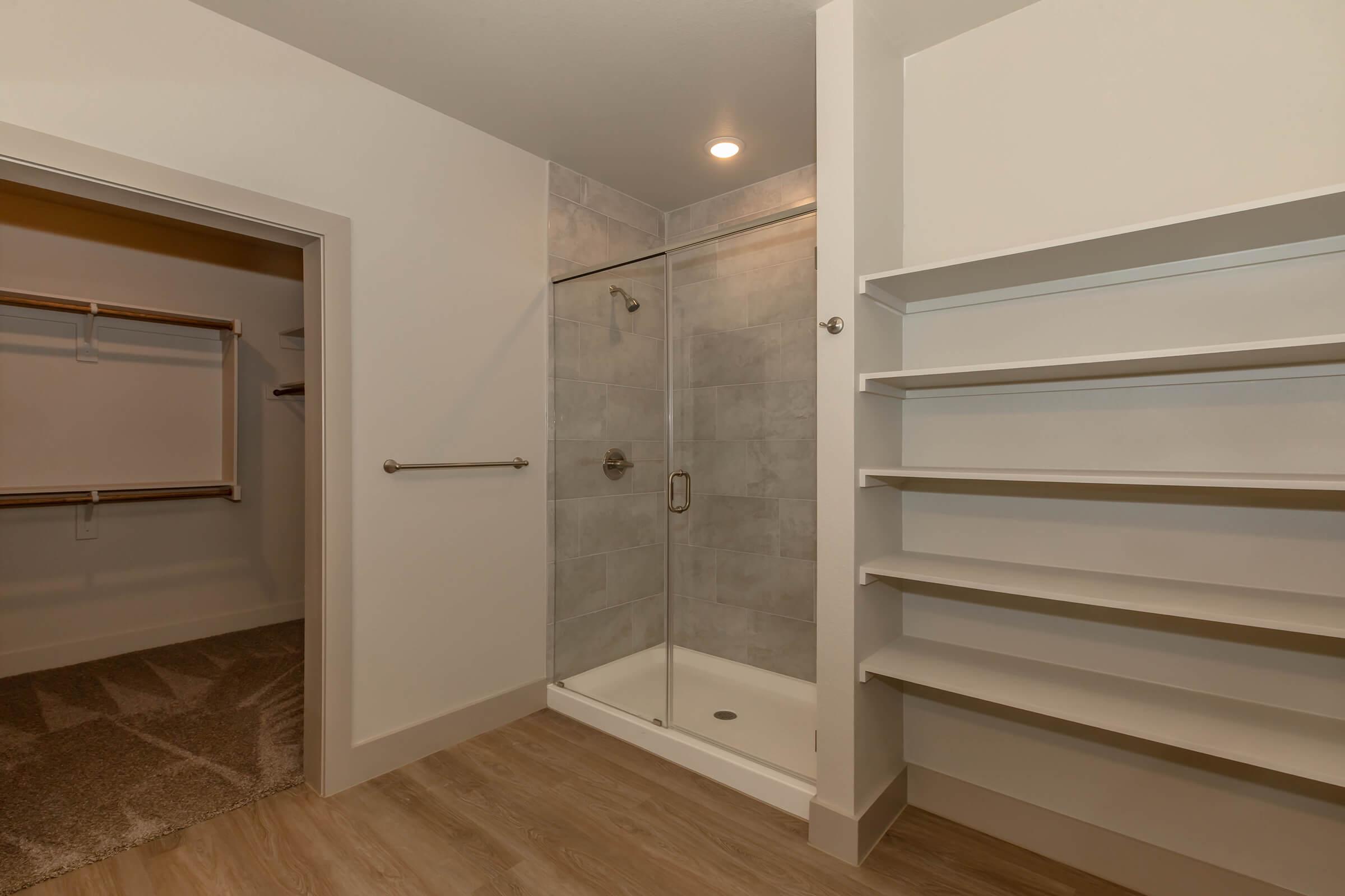 A modern bathroom featuring a glass-enclosed shower with gray tiled walls, a sleek shower fixture, and a white shower base. To the left, there is a walk-in closet with an open clothing rack, and on the right, multiple white shelves for storage or display. The floor is light-colored wood.