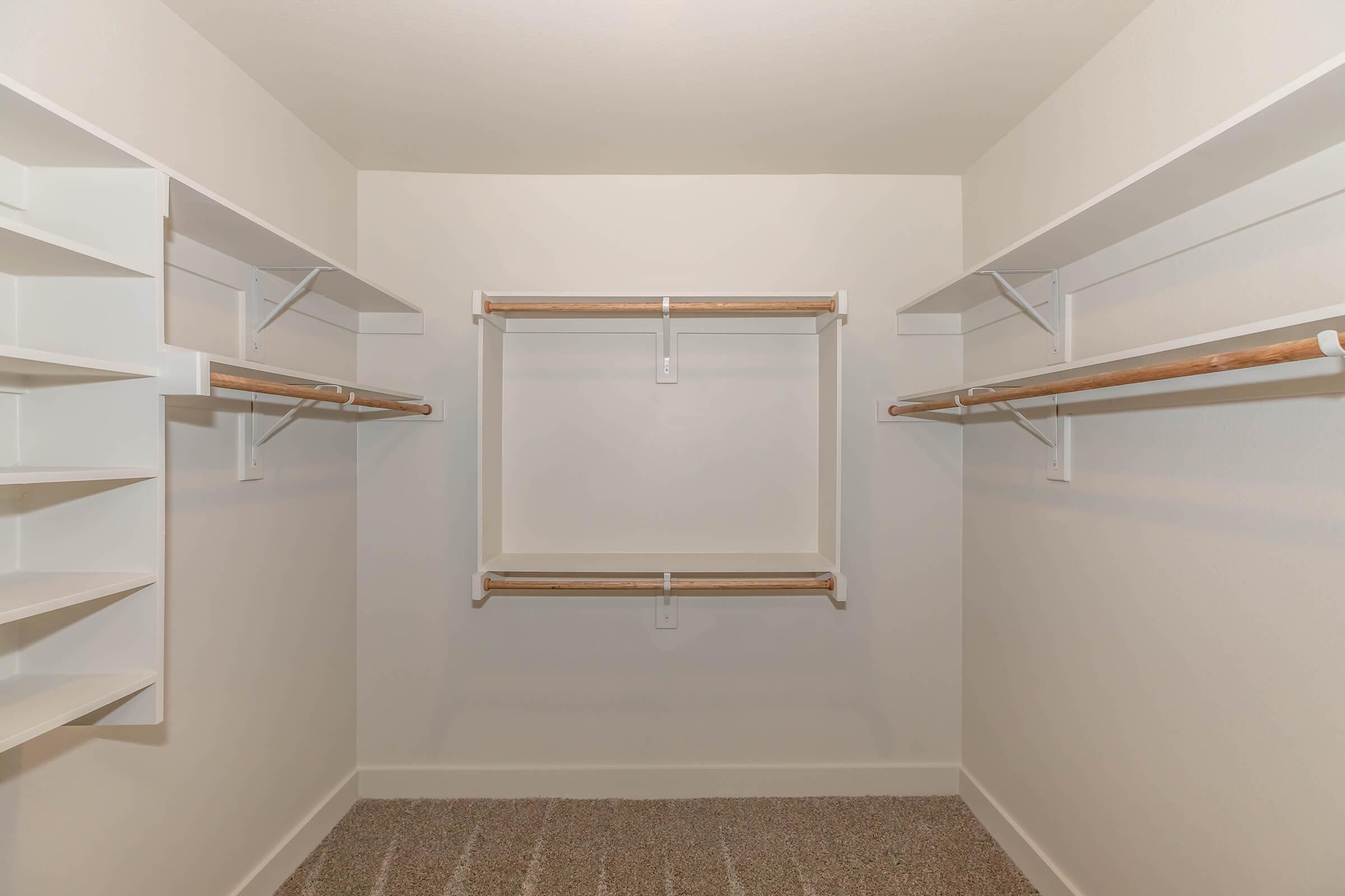 A spacious empty closet featuring white walls and shelving on the left side. There are two wooden hanging rods positioned at different heights, providing ample storage space. The floor is carpeted, and the overall design is simple and clean, allowing for organizational flexibility.