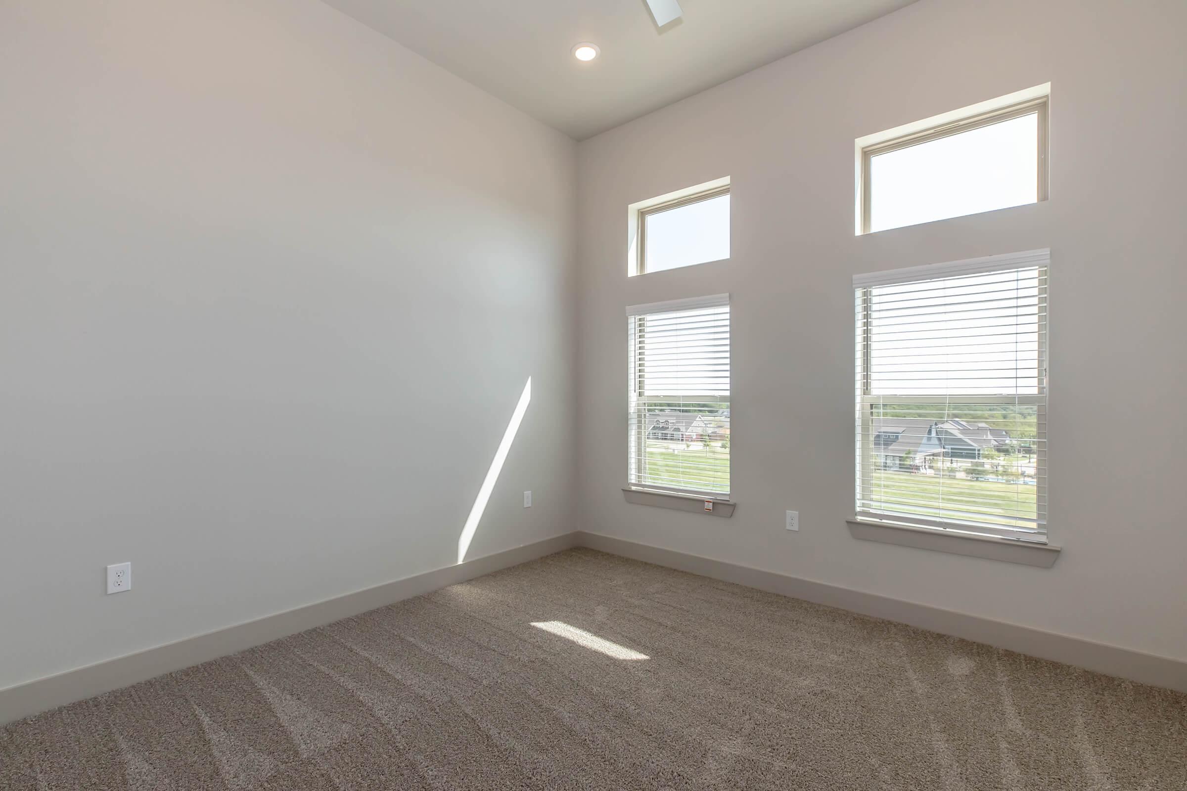 A bright, minimalist room with light gray walls and carpeted flooring. Two small windows are positioned on one wall, allowing natural light to enter. The room appears spacious and modern, featuring sloped ceilings and a clean aesthetic, suitable for various uses like an office or guest room.