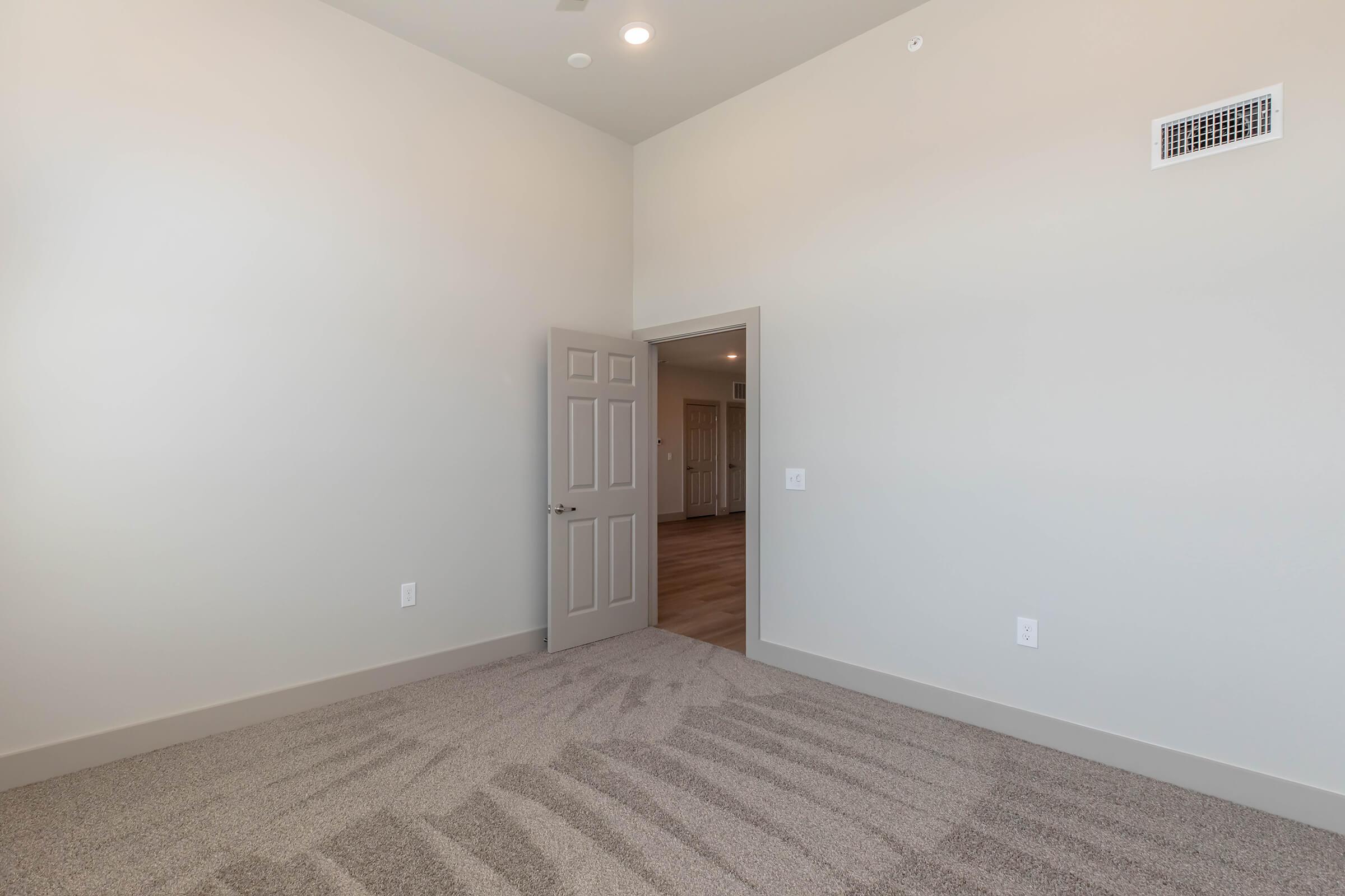 A spacious, empty room with light-colored walls and carpeted flooring. A slightly open door leads to another room, and ceiling lights are visible. The room has a neutral color scheme, providing a bright and airy atmosphere.