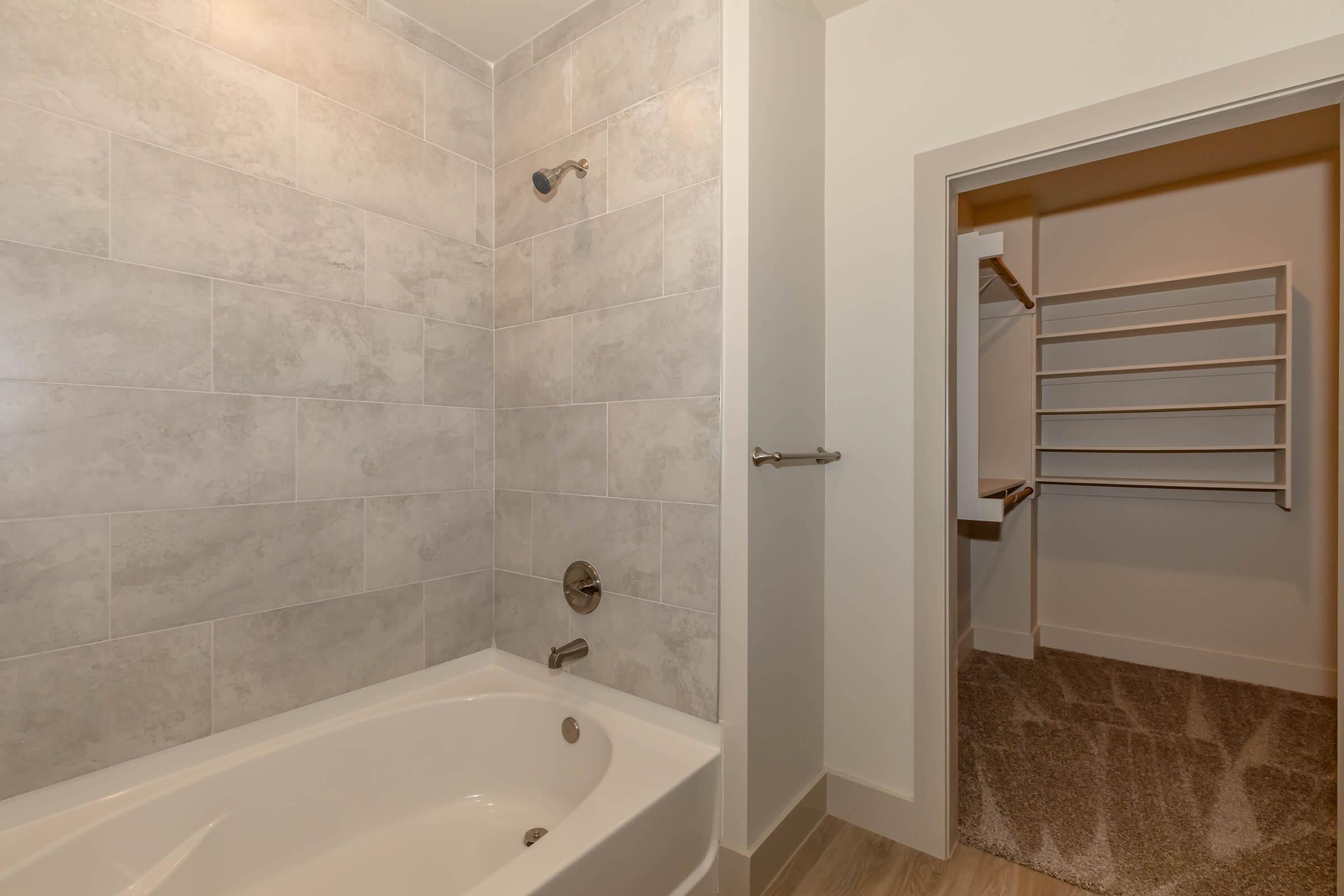 A modern bathroom featuring a tub with a shower, surrounded by light gray tile walls. To the right, there is an open closet with shelving. The floor is covered with a soft, light-colored carpet.