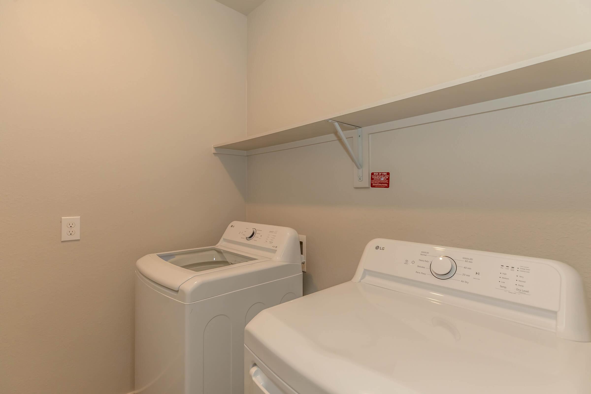 A laundry room featuring two white appliances: a washing machine and a dryer. The space includes a shelf above the machines and a wall with a small red warning sign. The walls are painted a light neutral color, creating a clean and functional environment for laundry tasks.